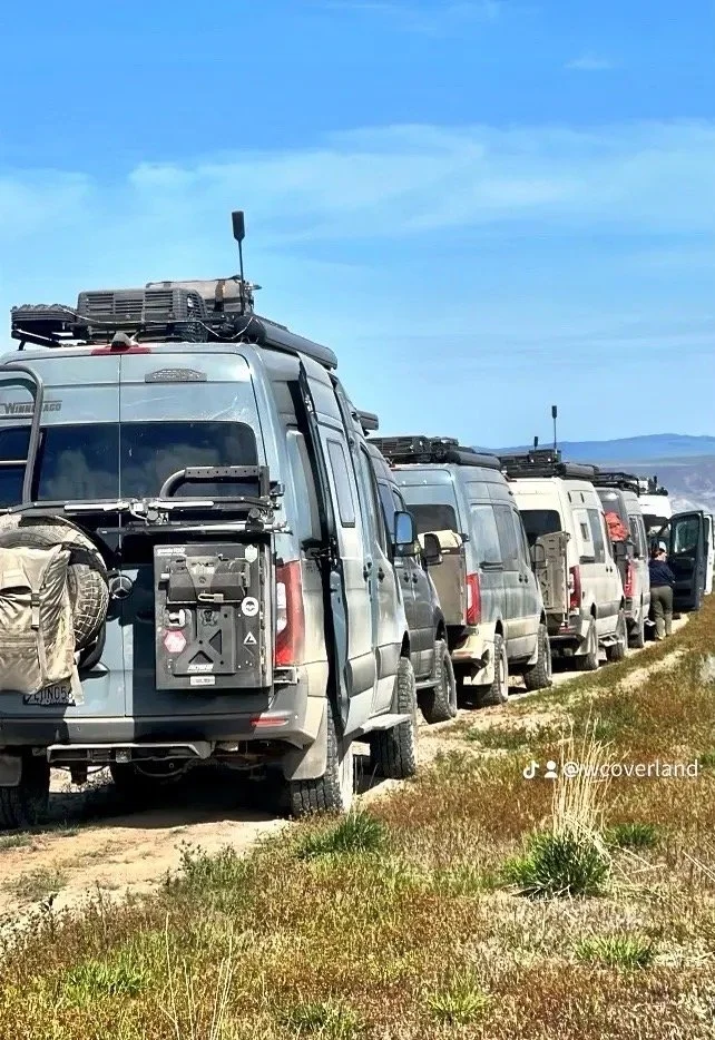 A line of off-road vehicles, mostly Mercedes-Benz Sprinter Vans, parked on a grassy dirt road under a blue sky.