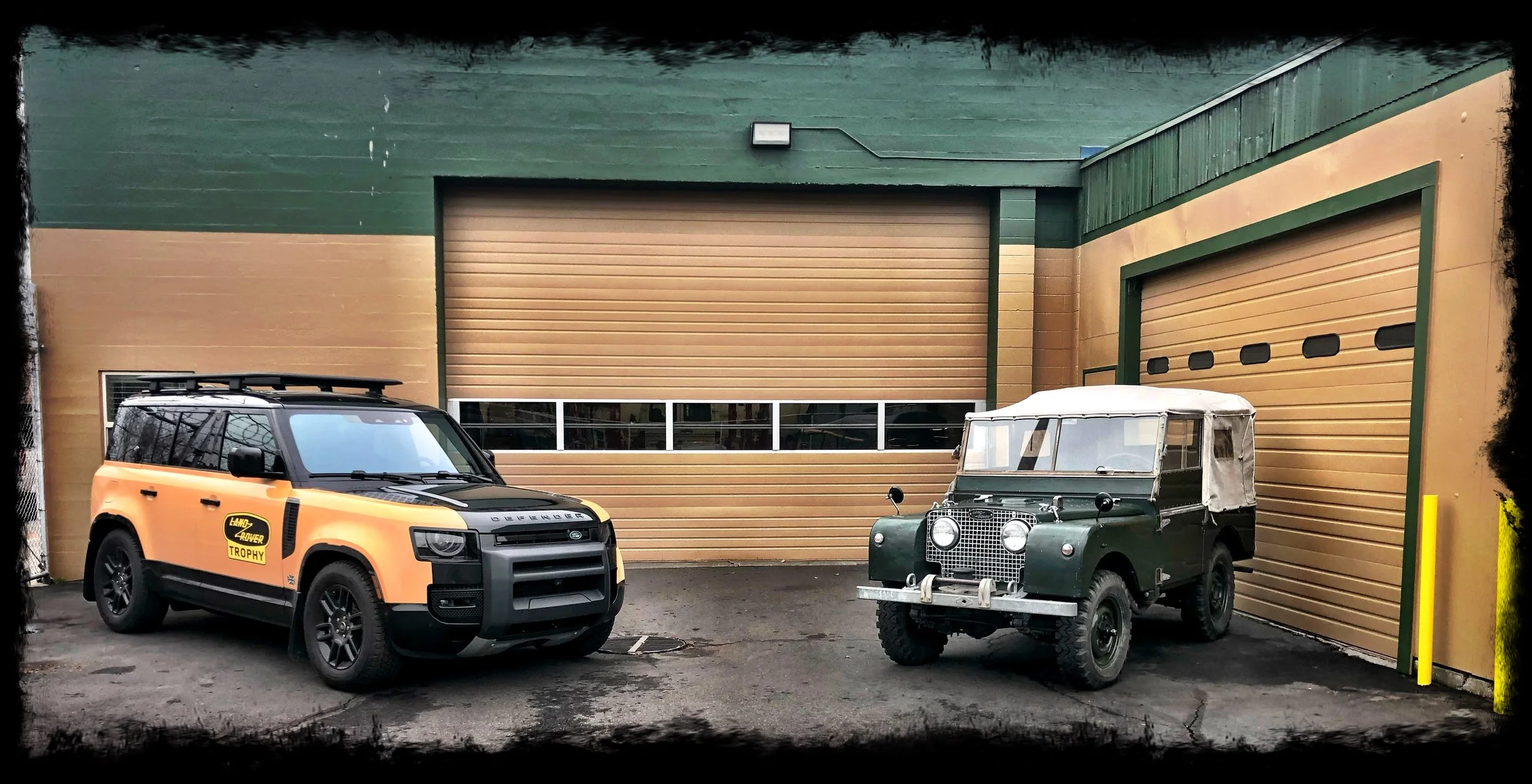 Two vintage and modern Land Rover vehicles parked in front of a large green and tan building with garage doors.