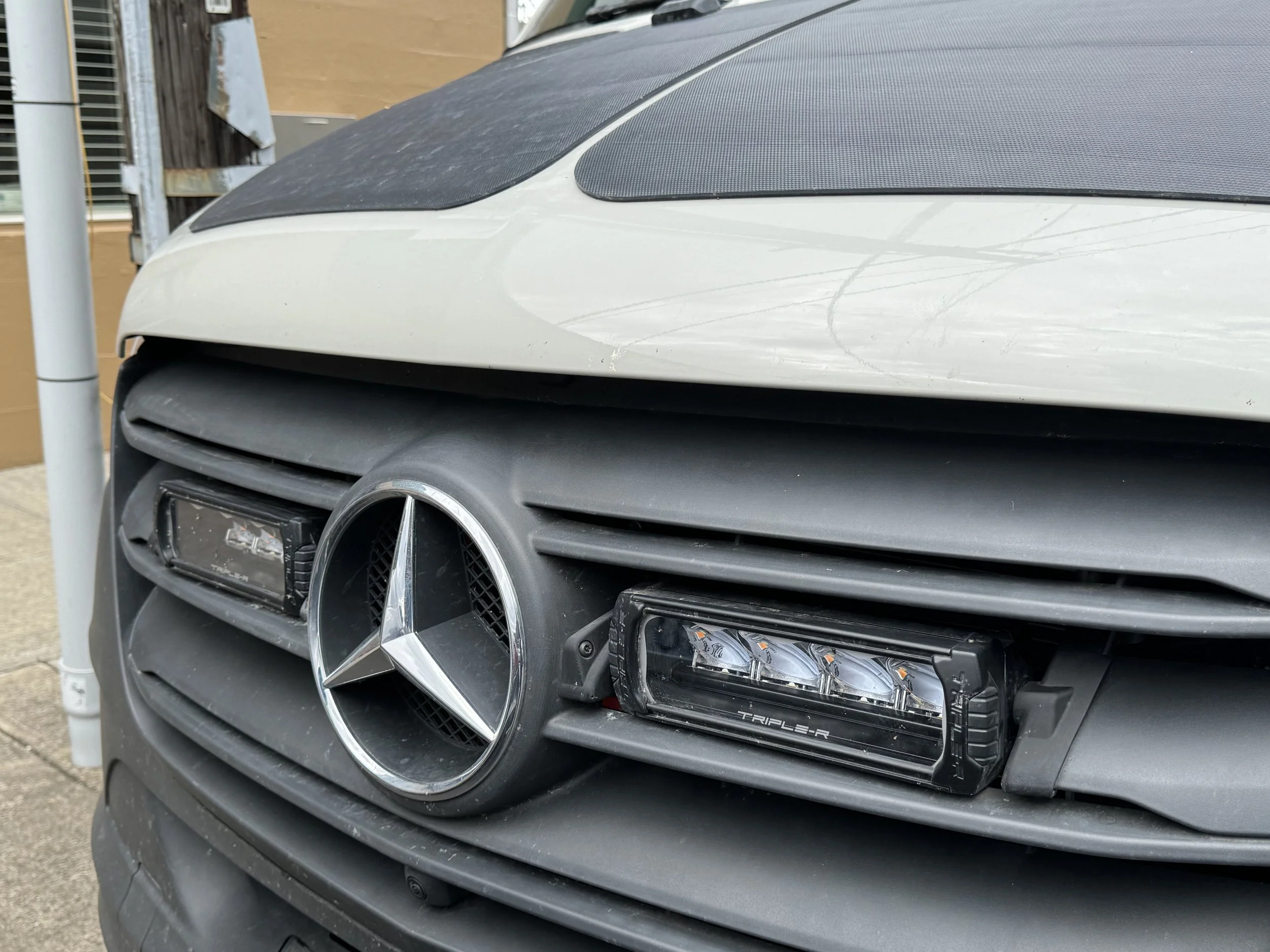Close-up of the front of a black Mercedes-Benz Sprinter van, showing the grille, one headlight, and a carbon fiber hood cover.