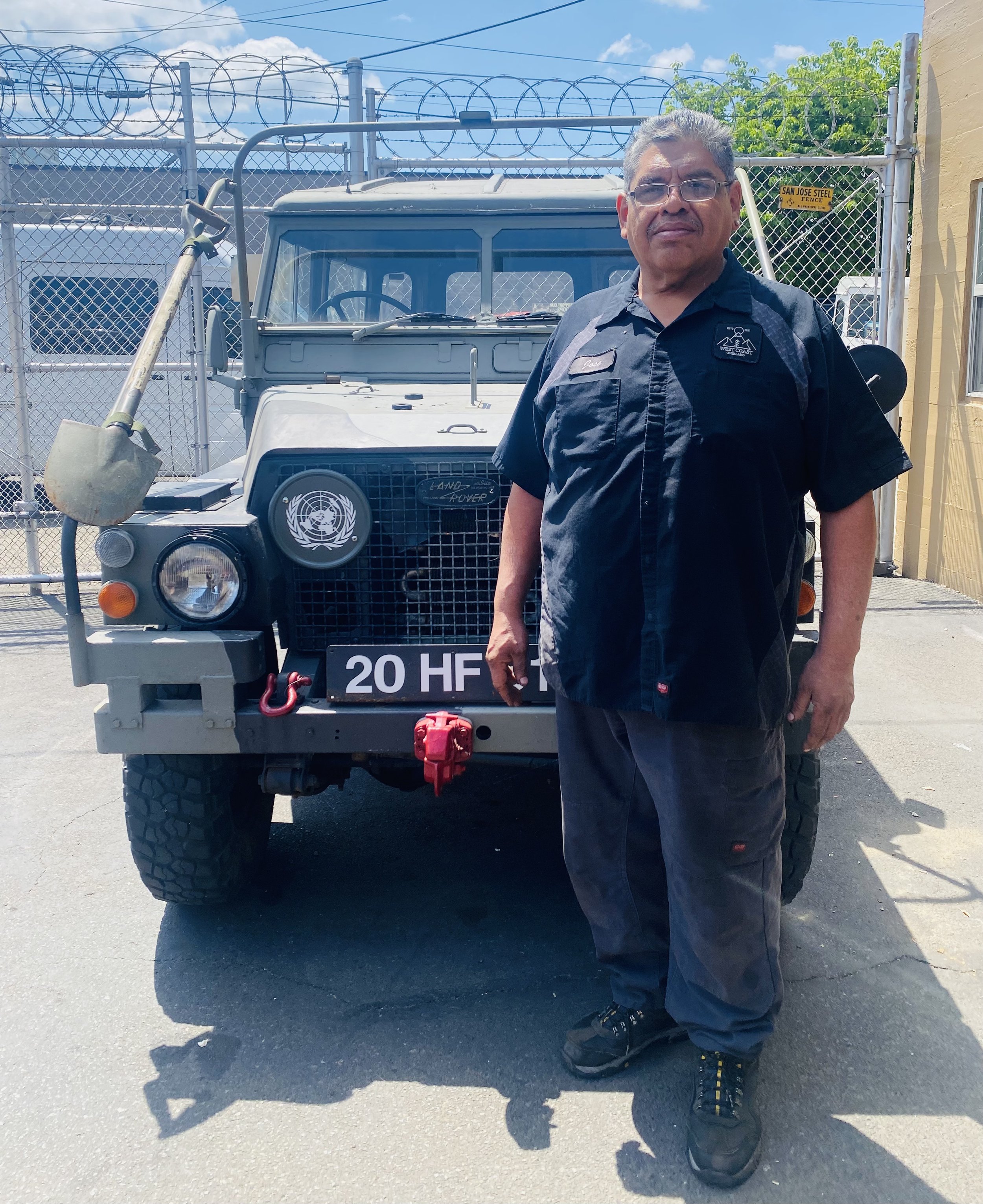 A man with glasses and gray hair kneeling on the ground next to a gray Land Rover Defender with a rack and a shovel on the hood. The background includes a chain-link fence topped with concertina wire, a yellow building on the right, and clear blue sky with some clouds.