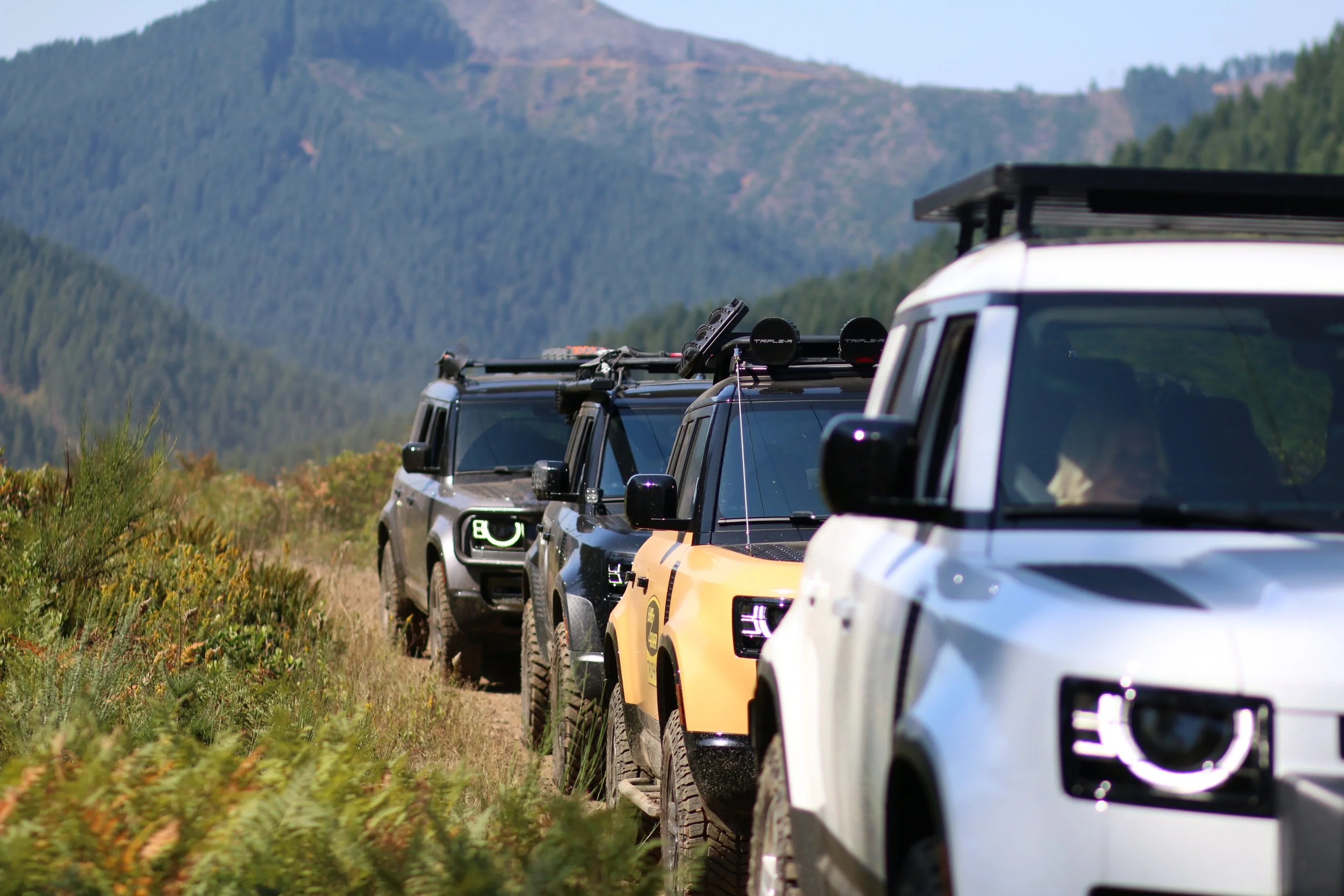 Line of four Land Rover Defender SUVs in an outdoor mountainous area, with hills and green trees in the background.