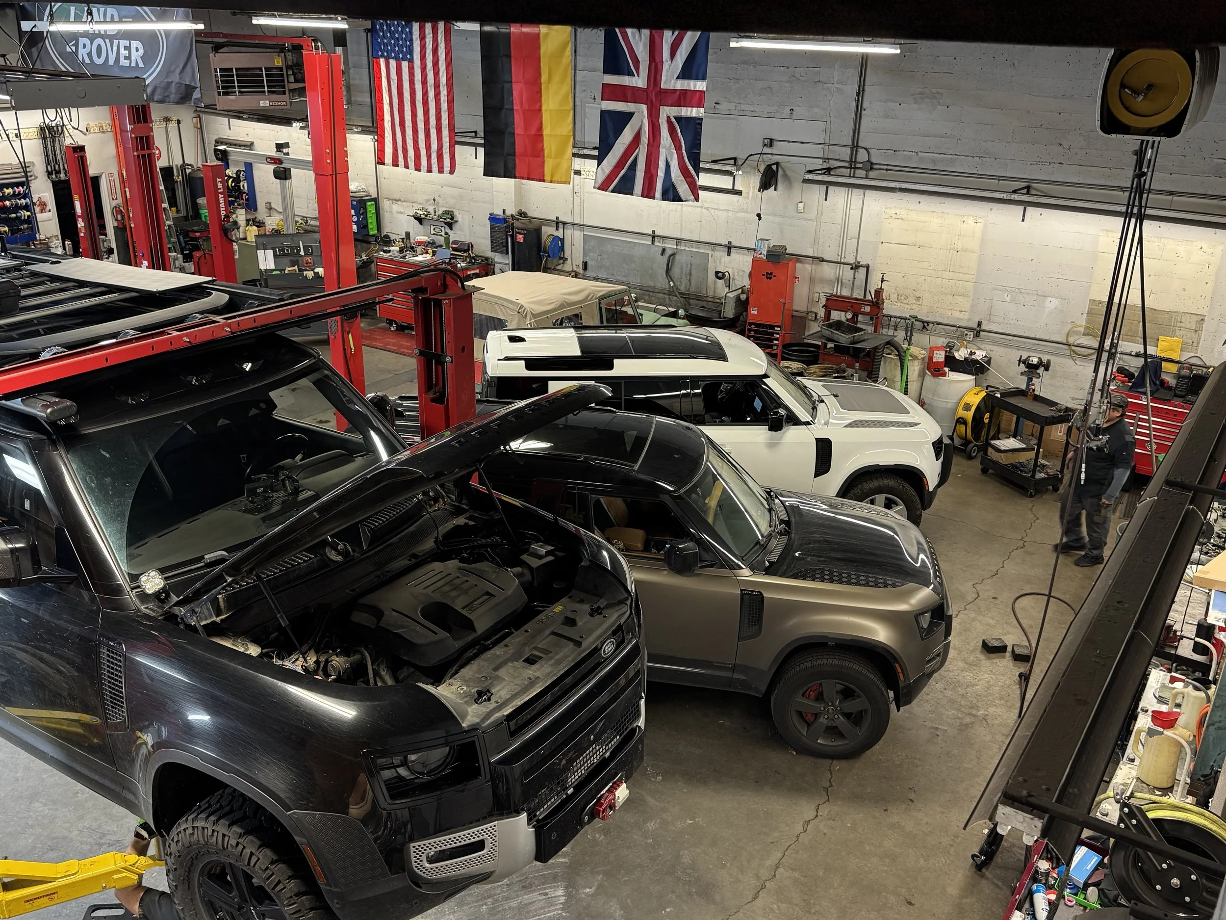 An auto repair shop with several luxury cars, including a black car, a white car, and a silver car, inside a concrete-floored garage. American, British, and German flags hang on the wall, and various tools and equipment are visible, including red too
