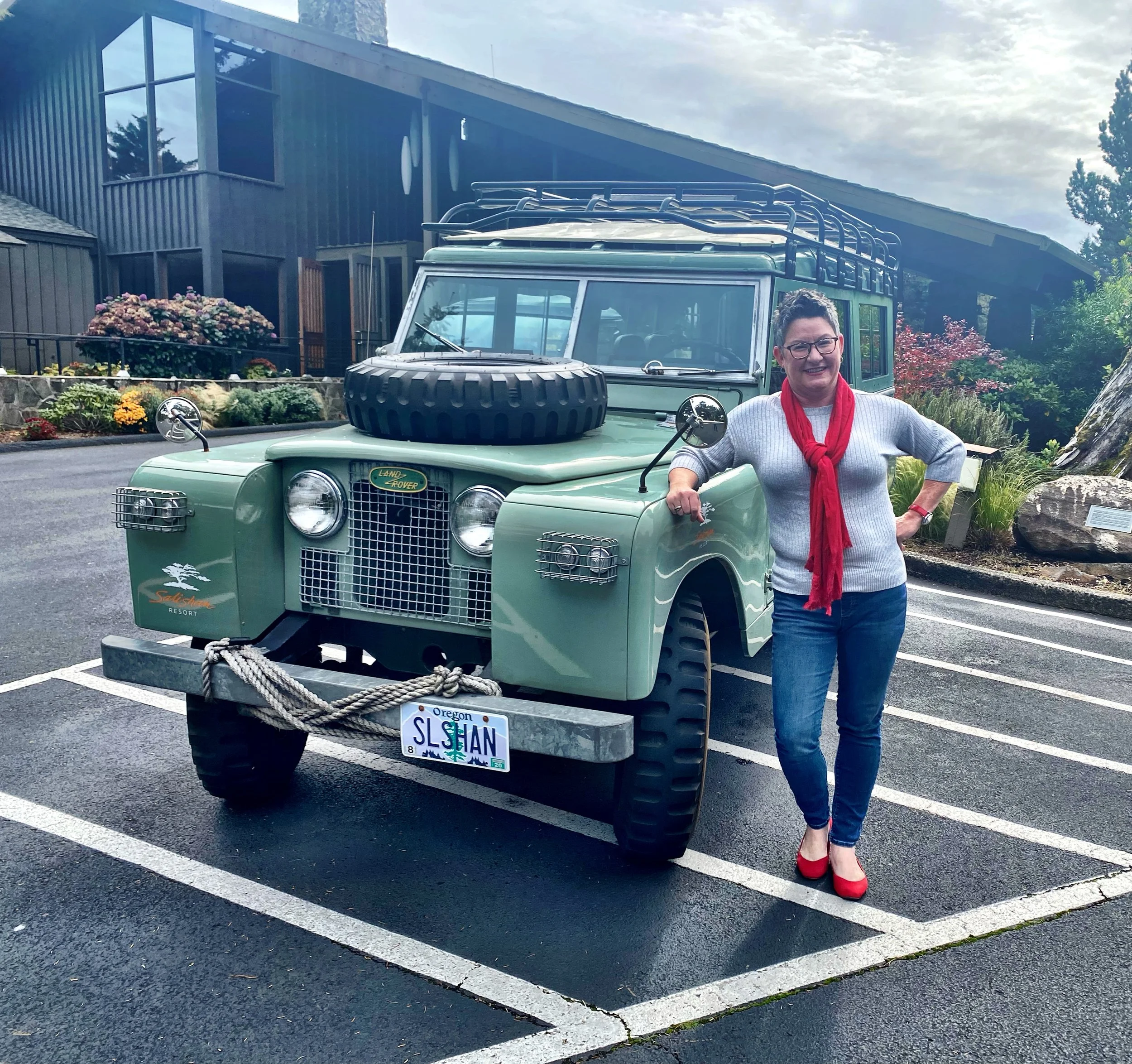 A woman standing next to a vintage Land Rover vehicle with a spare tire on the hood, in front of a modern building with garden flowers and trees, wearing glasses, a gray sweater, a red scarf, blue jeans, and red shoes.