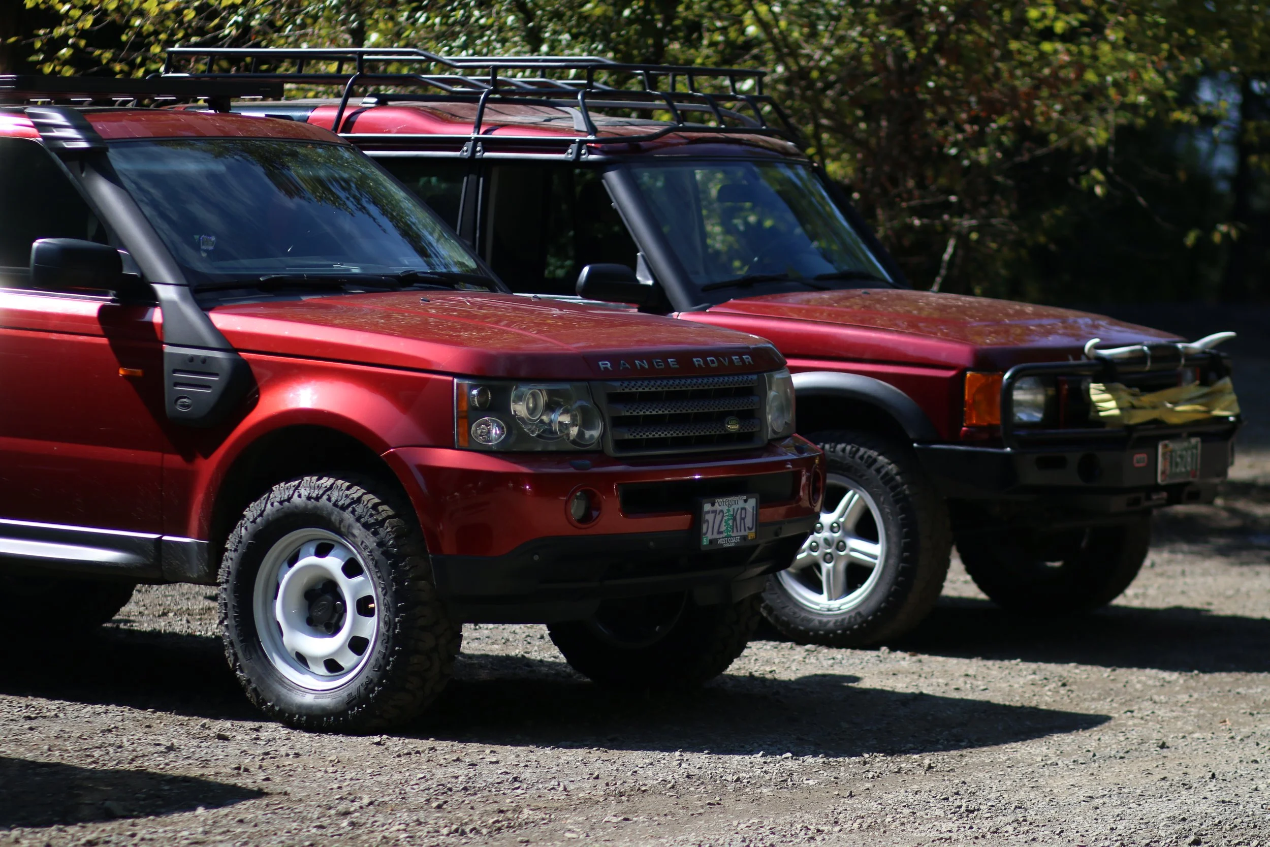 Two red Land Rover SUVs parked on a dirt ground with trees in the background.