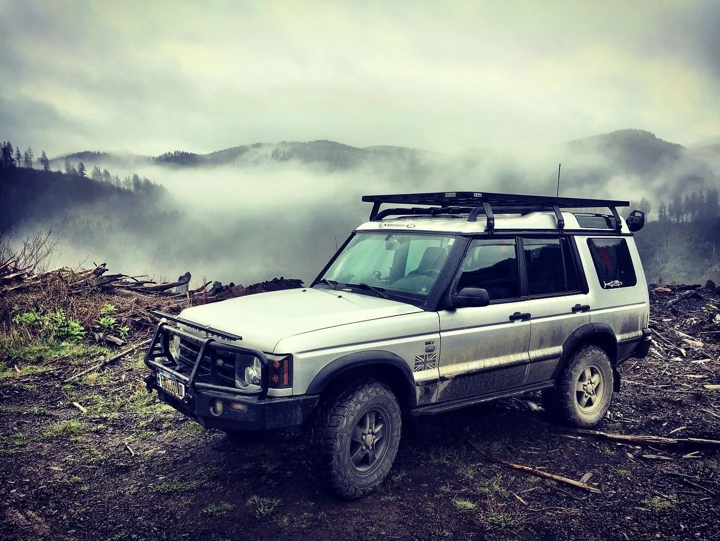 An off-road vehicle parked on muddy ground with a misty mountain landscape in the background.