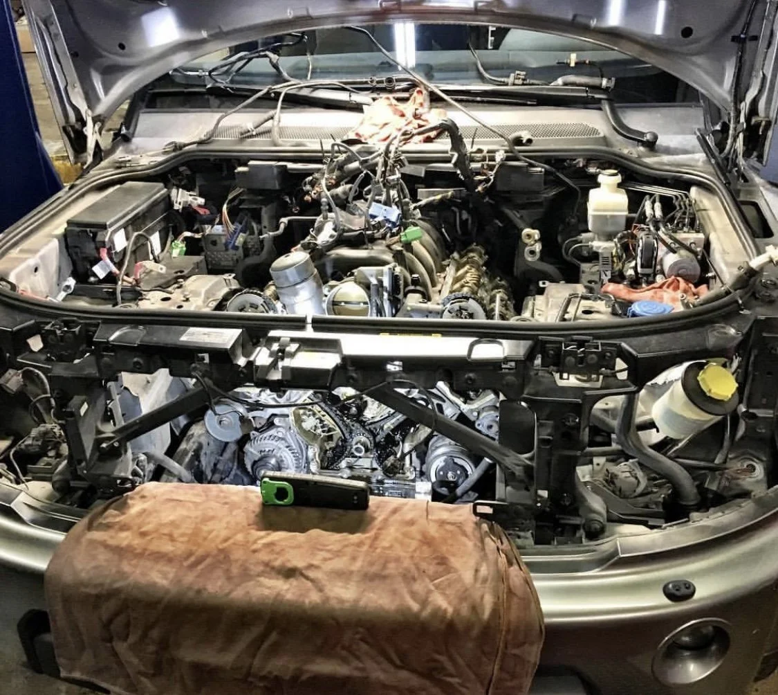 Land Rover engine bay with work in progress, engine parts exposed, tools and fluid reservoirs visible, and a mobile device on a cloth in the foreground.