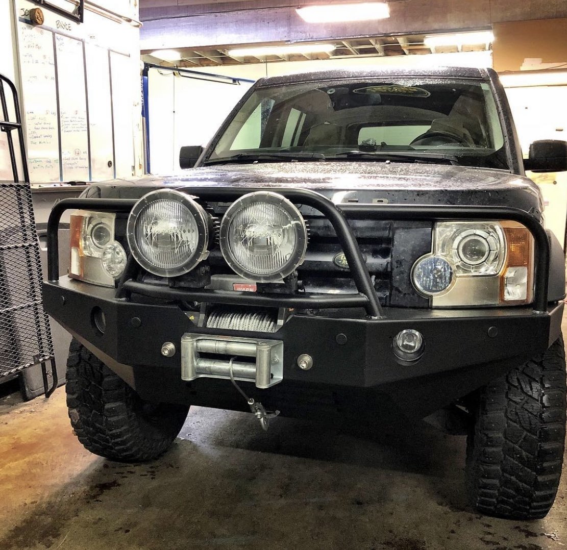 Front view of a black Land Rover LR3 with large tires, a heavy-duty bumper, and four large round headlights, parked inside a garage or workshop.