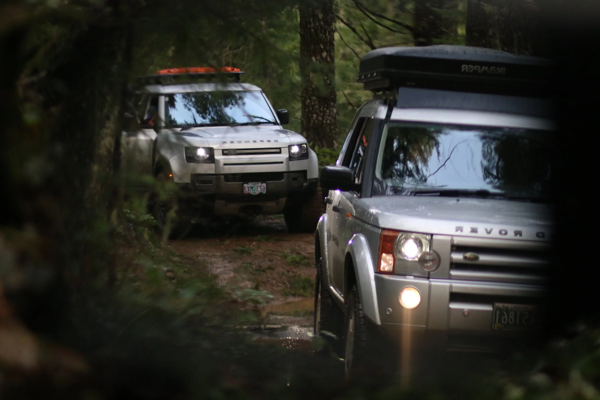 Three Land Rover off-road vehicles driving on a wet forest trail surrounded by trees, with the view partially obscured by foliage.