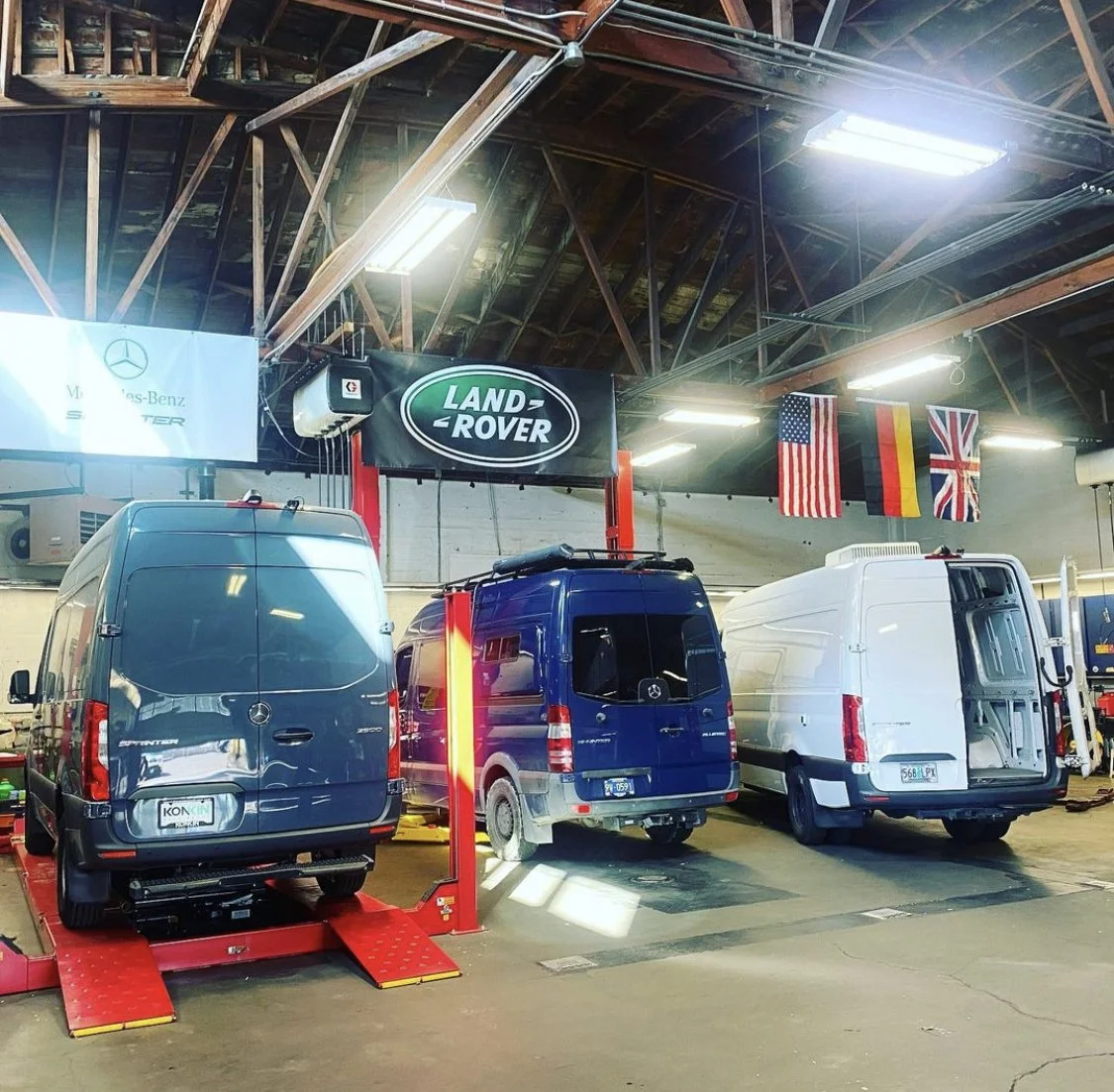 Indoor auto repair shop with three Mercedes-Benz vans on hydraulic lifts, flags of the United States, Germany, and United Kingdom hanging from the ceiling, and signs for Land Rover and Mercedes-Benz.