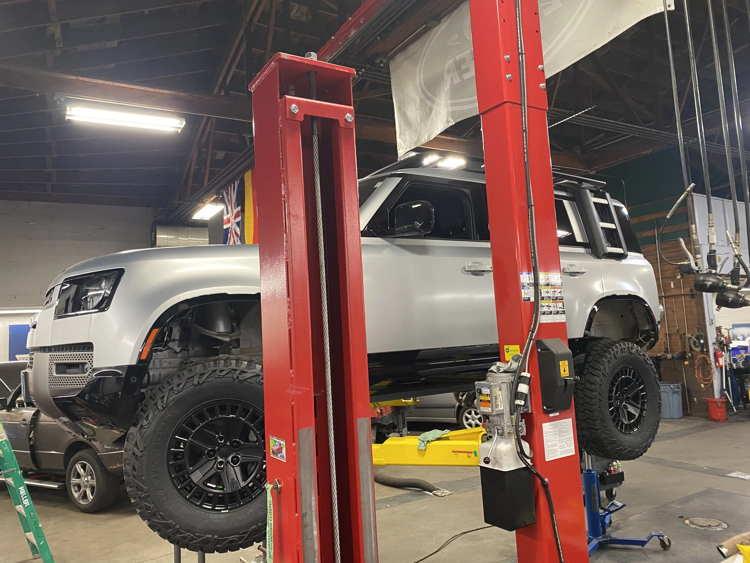 A silver Land Rover is lifted on a red hydraulic automotive lift inside a garage or workshop. There are tools and equipment visible around the workspace.