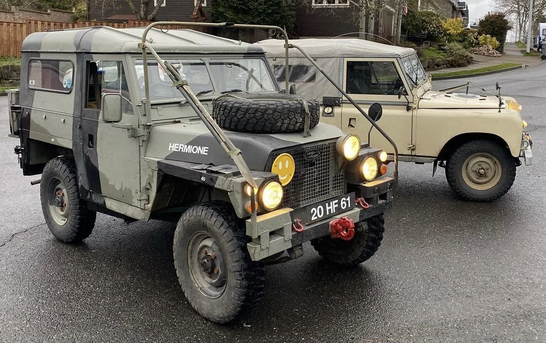 Two vintage off-road vehicles parked on a wet street. The vehicle in the foreground is painted in camouflage, has large tires, a spare tire on the hood, and a yellow smiley face sticker on the front grille. The vehicle in the background is beige with