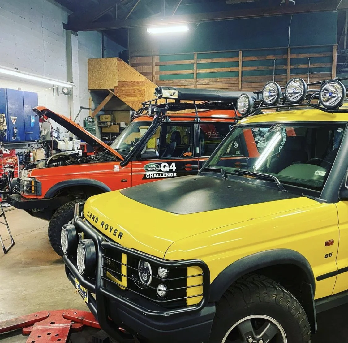 Two vintage Land Rover vehicles inside a garage, one yellow with a black hood and a front bumper with multiple lights, and one red with a black roof rack and an open hood, surrounded by tools and workshop equipment.