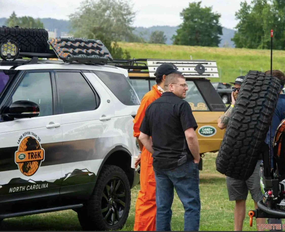 Group of people standing outdoors beside Land Rover vehicles equipped for off-road adventure, with some carrying spare tires and gear.