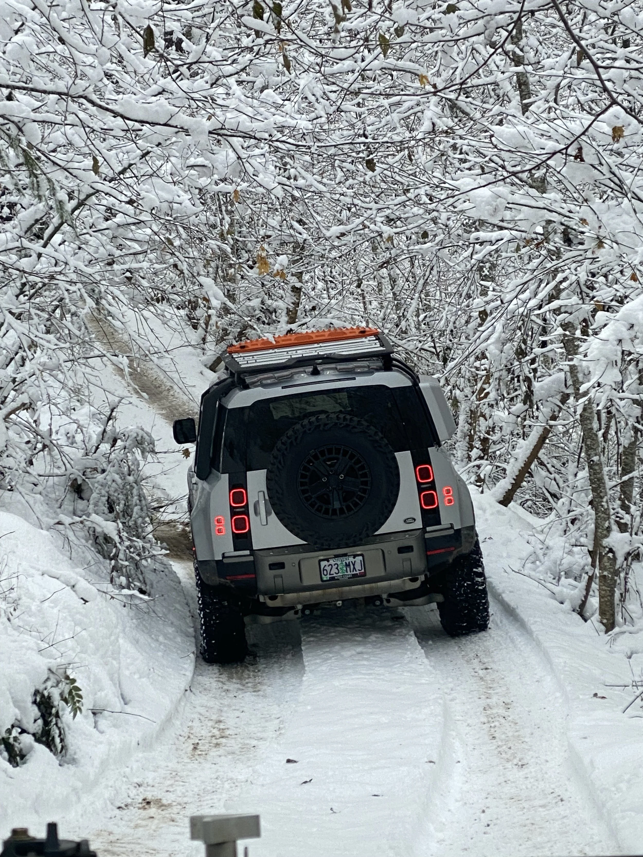 A Land Rover Defender driving on a snow-covered trail through a dense winter forest with snow-laden branches.