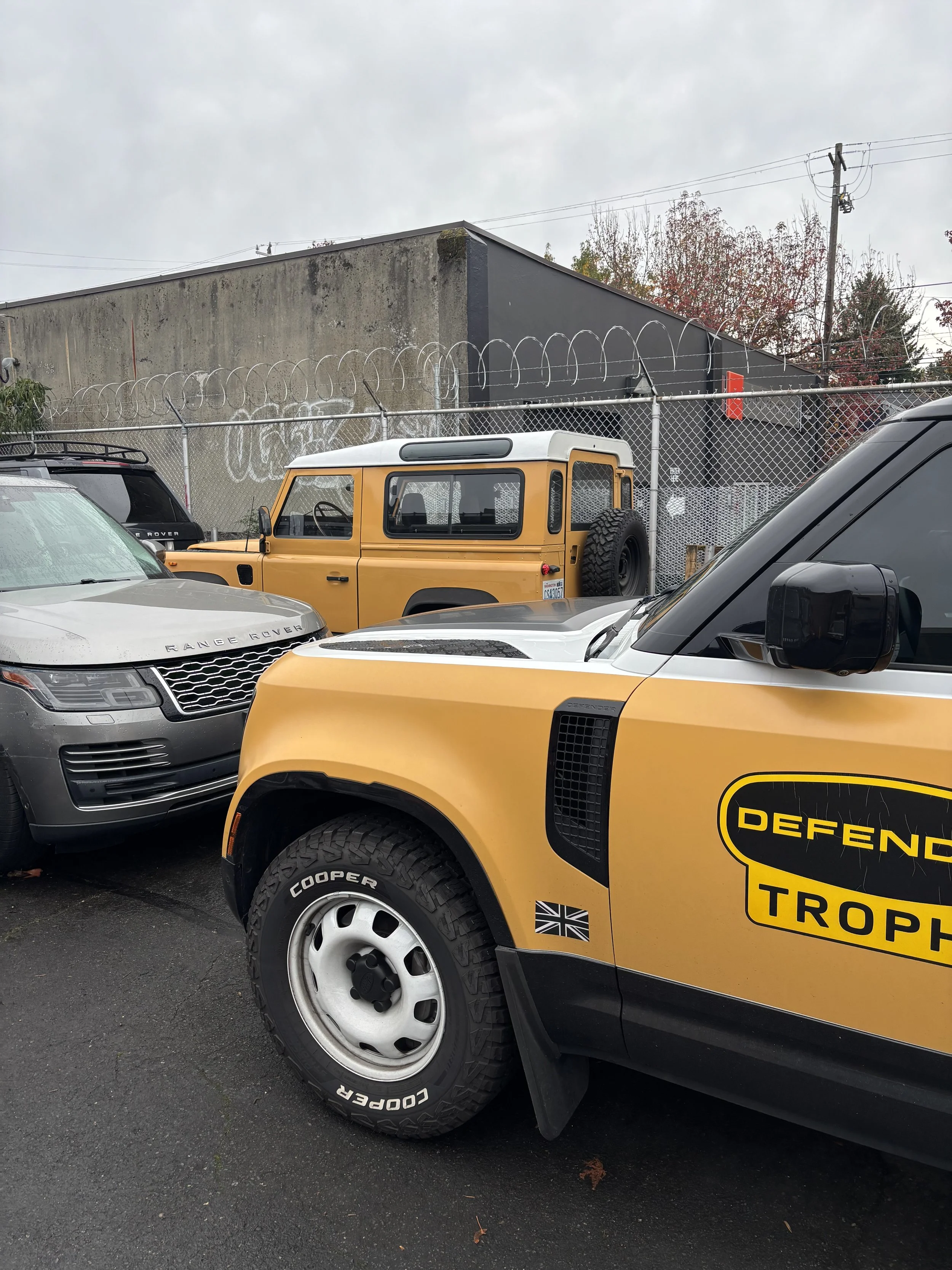 A parking lot with multiple vehicles including a gray Range Rover, a yellow Defender troop vehicle, and parts of other cars. The background shows a chain-link fence topped with barbed wire and a weathered concrete building with graffiti, under a clou