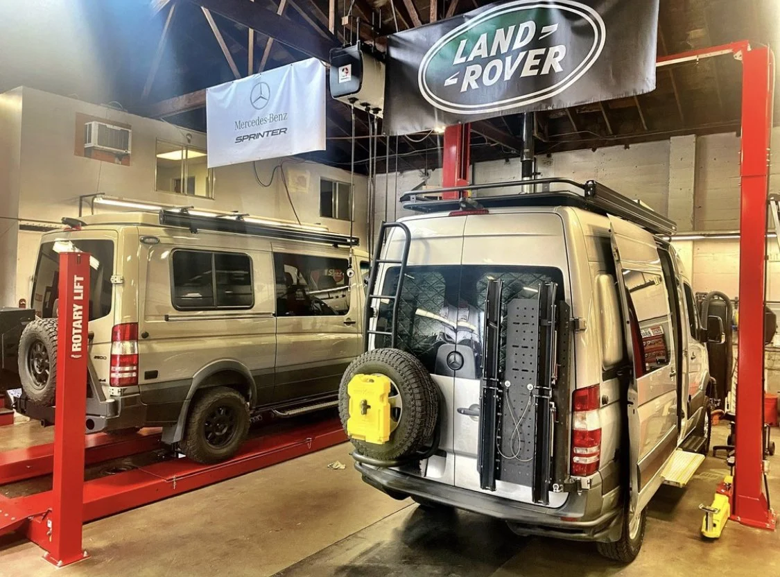 Two beige Mercedes-Benz Sprinter vans inside a garage or workshop, one on a vehicle lift with a roof rack and spare tire, the other parked next to it with a similar roof rack and equipment attached to the rear. There are banners with Land Rover and M