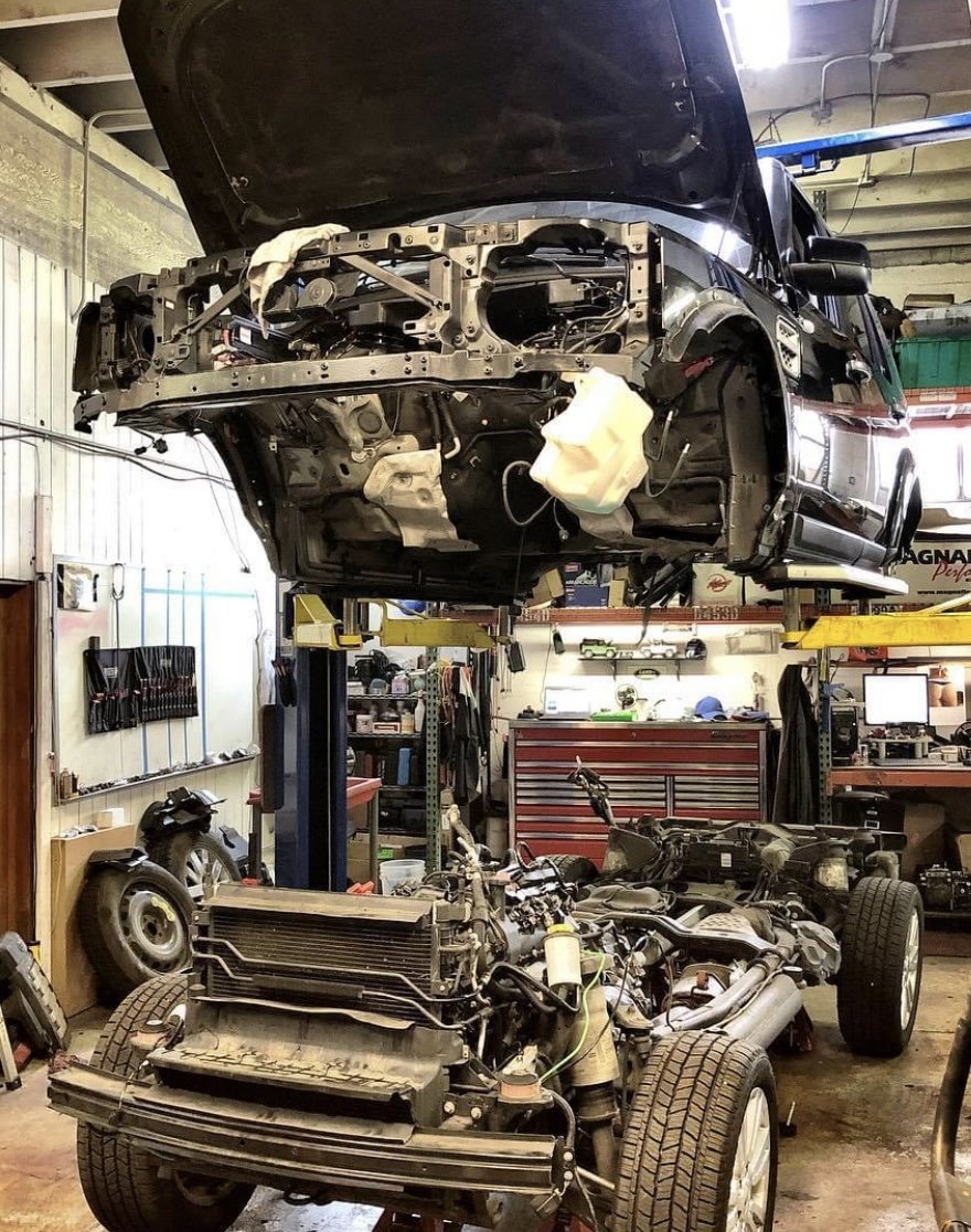 A Land Rover with its hood open is elevated on a lift, exposing the engine compartment. Below, the car's body is disassembled, revealing internal parts and frame, in a cluttered auto repair shop with tools, equipment, and workbenches.