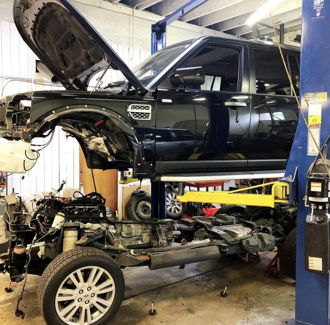 Black Land Rover on a hydraulic lift with the hood open, in an auto repair shop. The truck's front is disassembled, showing engine components and wheel well.