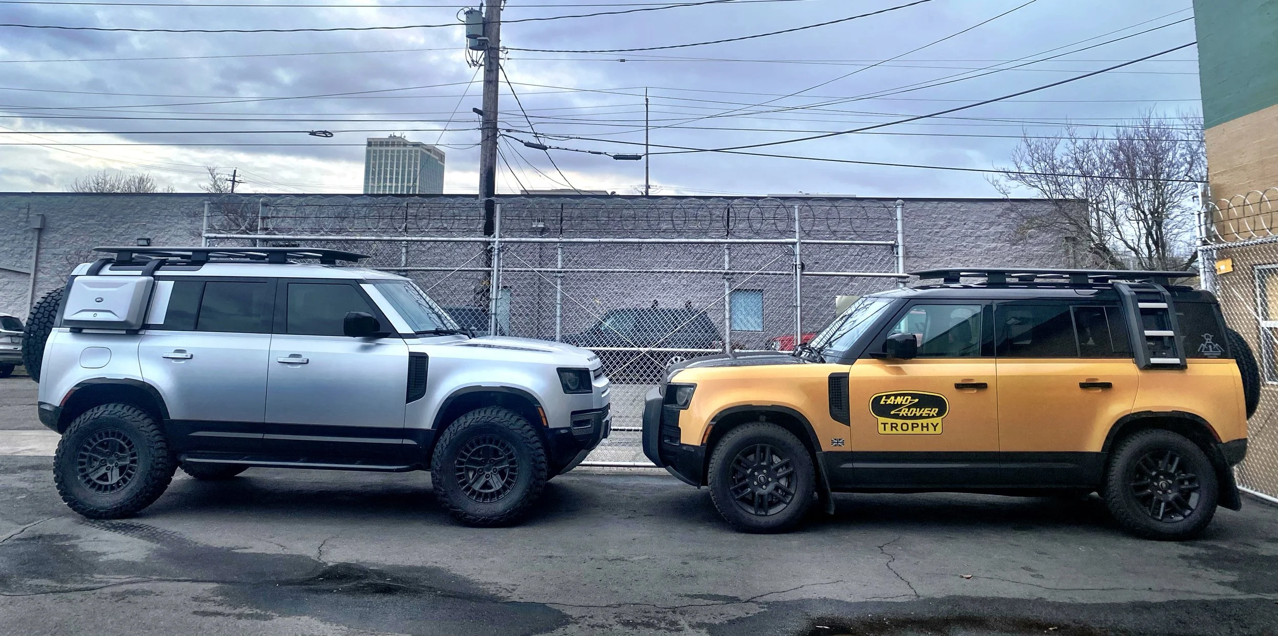 Two Land Rover vehicles parked side by side in an urban setting behind a chain-link fence with barbed wire on top, with overcast sky and power lines overhead.
