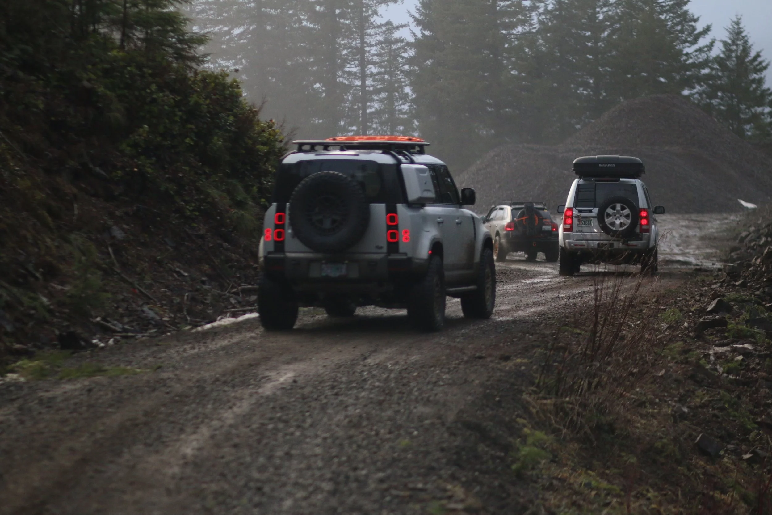 A convoy of four Land Rover off-road vehicles driving on a muddy forest trail with tall trees and mist in the background.