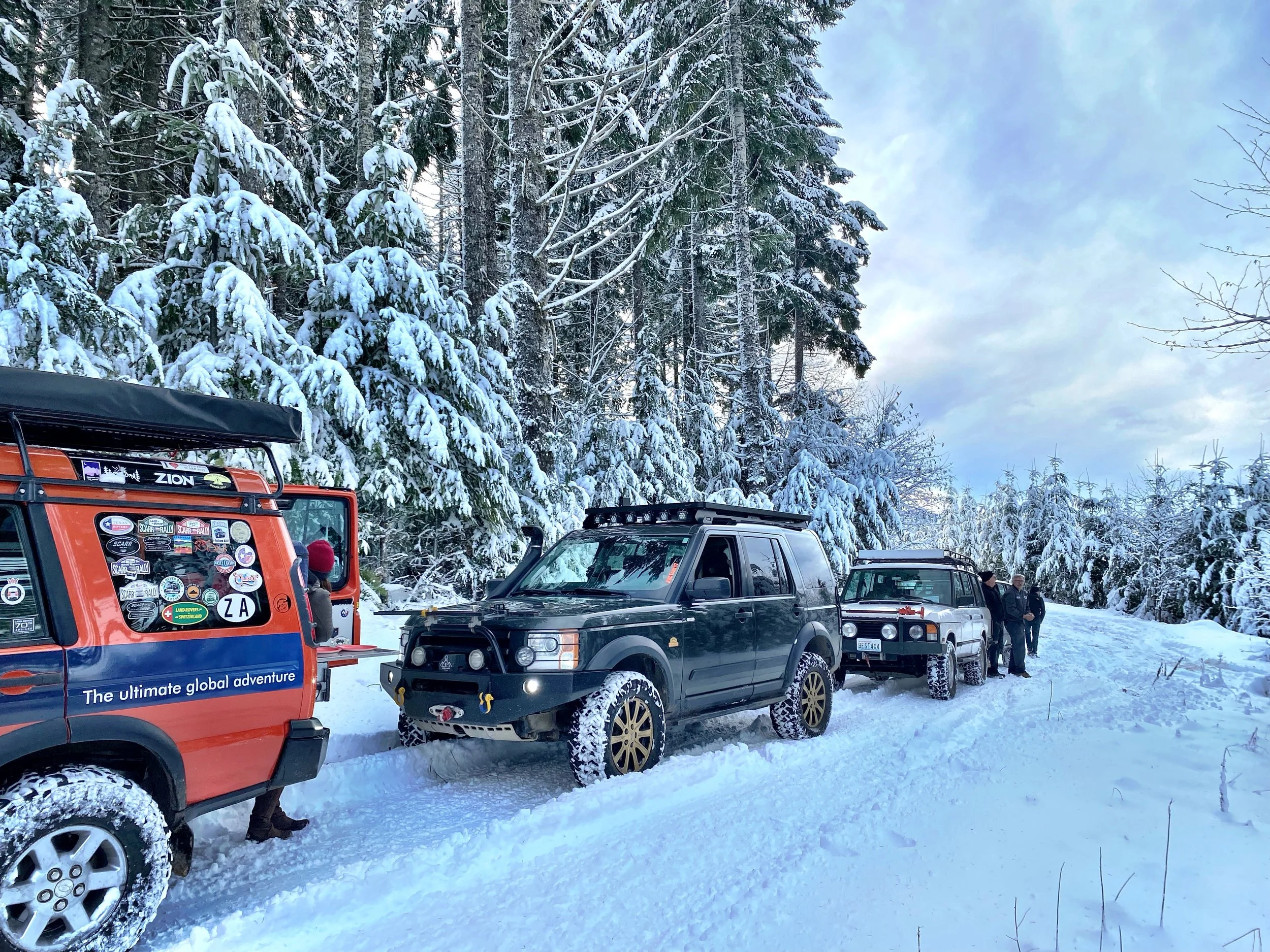 Snow-covered forest with several Land Rover SUVs parked off-road vehicles and a group of people standing in the snow.