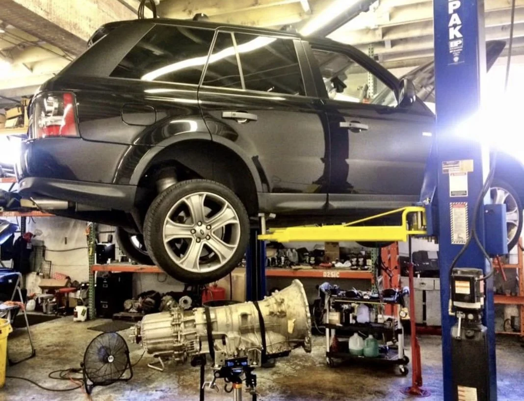 A black Range Rover Sport SUV is lifted on a car lift in an auto repair shop, with its wheels removed. The shop contains various tools, equipment, and a large engine transmission unit on the floor below.