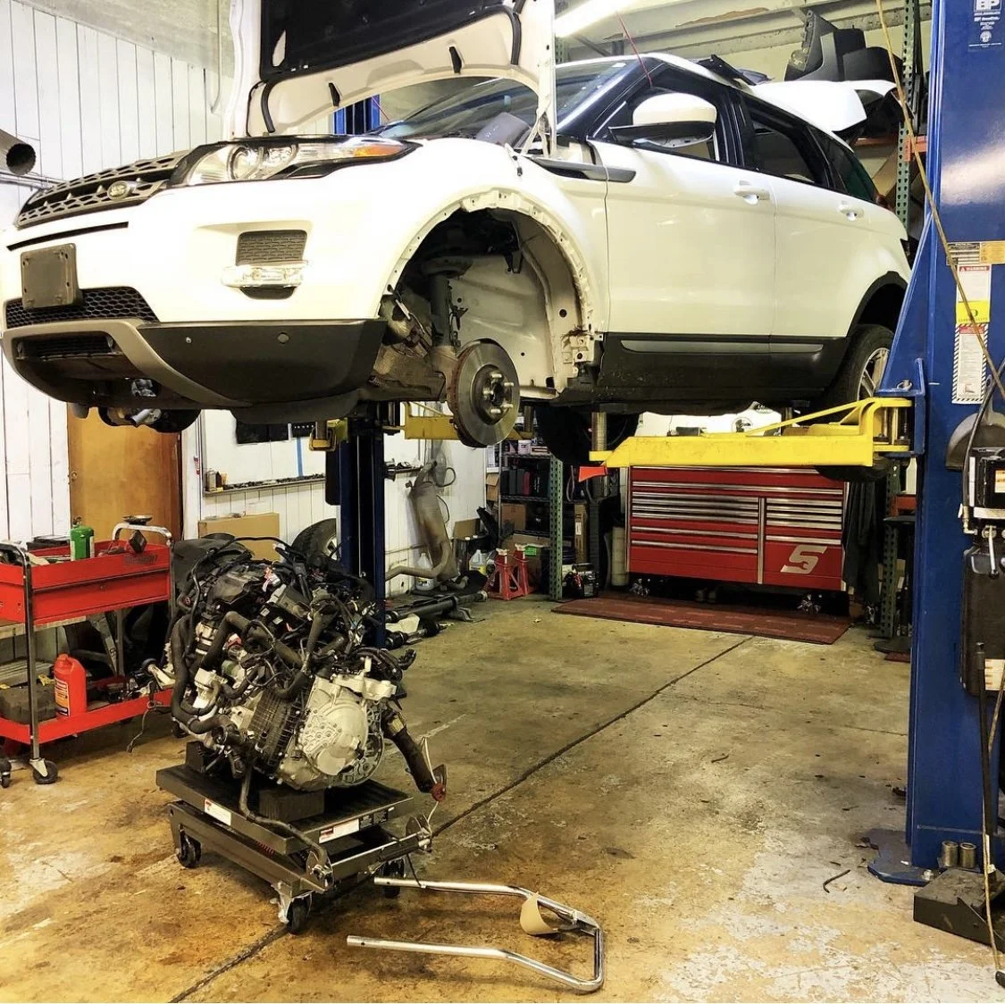 Auto repair shop with a white Range Rover SUV elevated on a lift, missing its wheels, and an engine on a stand nearby.