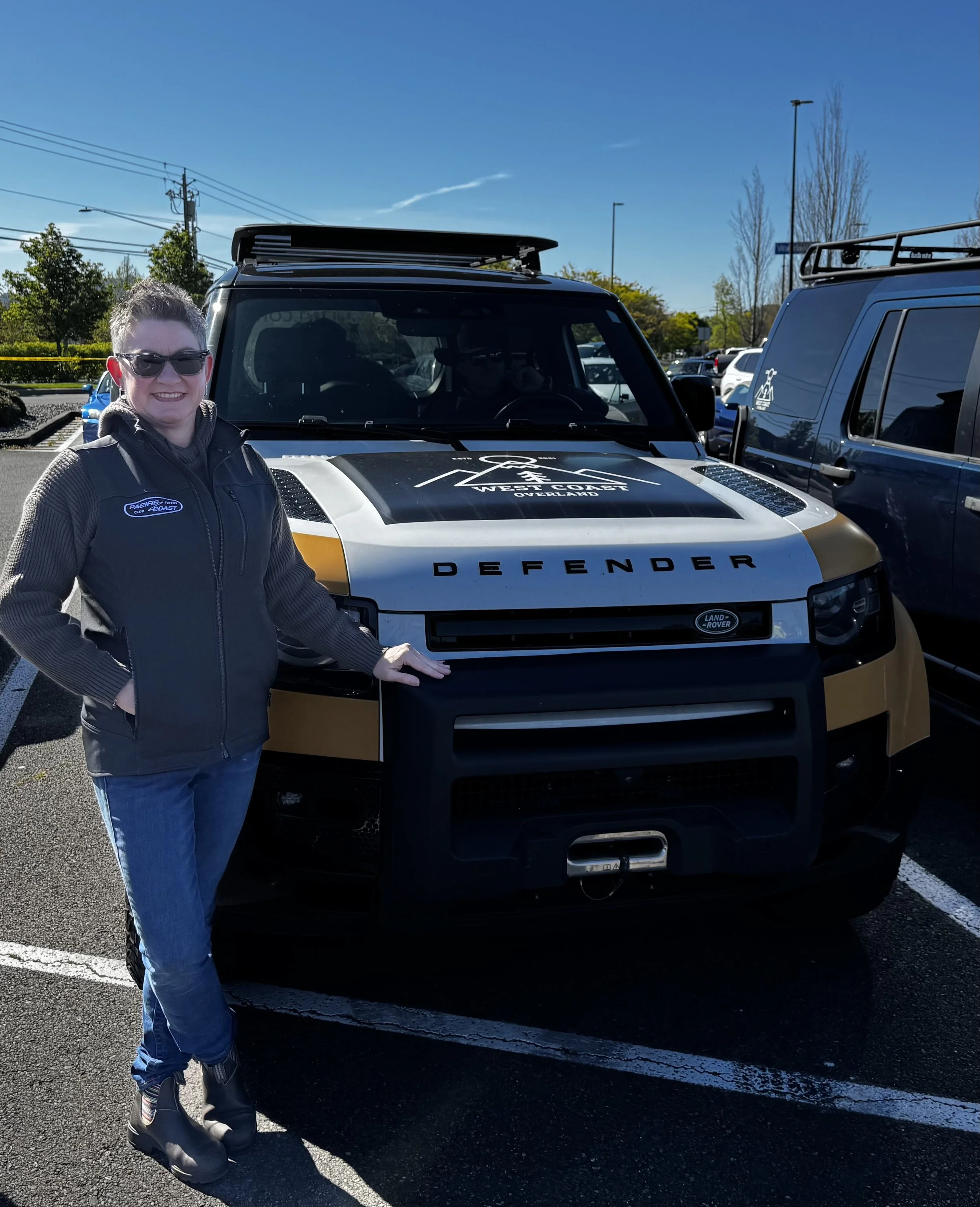 A woman wearing sunglasses and a black jacket with a patch that says 'Pacific Coast' is standing in a parking lot next to a black and white Land Rover Defender SUV with a roof rack. The vehicle has a logo on the hood that reads 'West Coast Overland.'