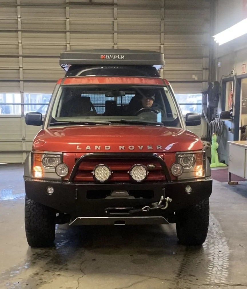 Red Land Rover parked inside a garage with a black roof cargo box and off-road lights on the front bumper.