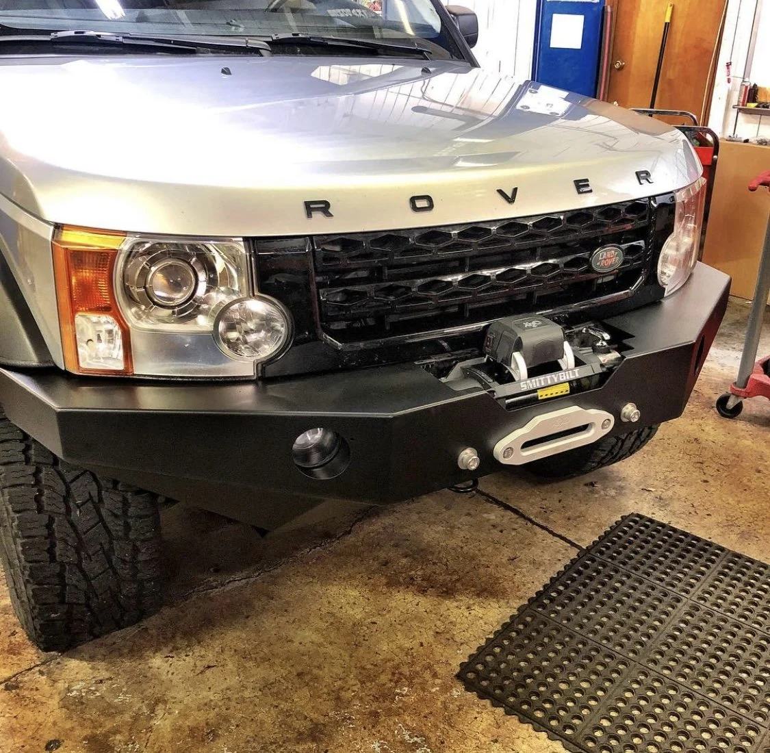 Front of a silver Land Rover vehicle with a black aftermarket bumper and winch in a garage.