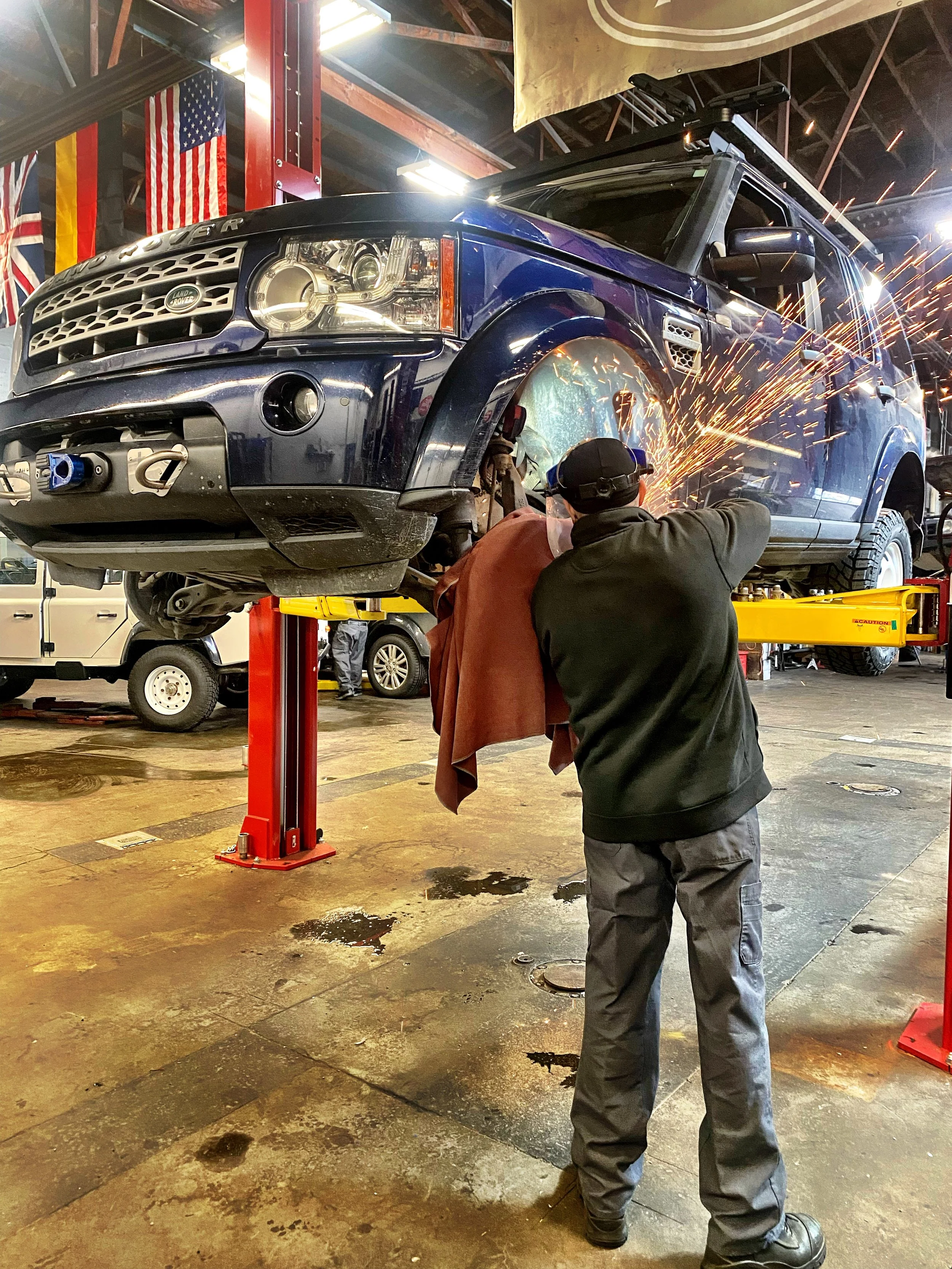 Two mechanics working on a blue Land Rover Discovery vehicle elevated on a hydraulic lift in an auto repair shop, with one mechanic welding and sparks flying, and American flags hanging in the background.
