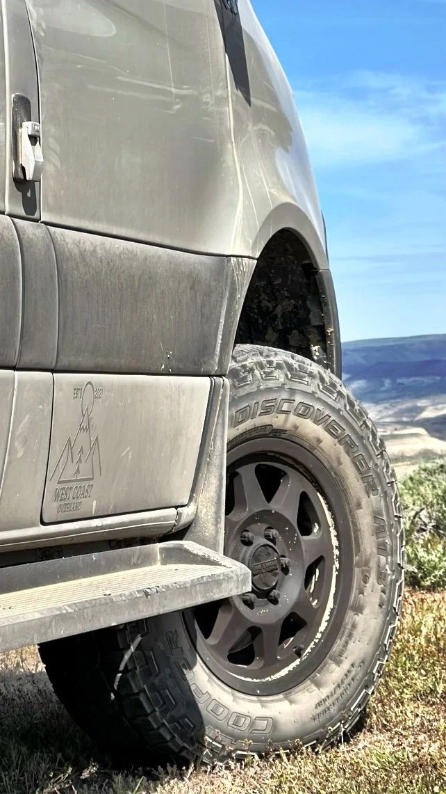 Close-up of a dirty Sprinter Van tire with rugged tread, mounted on a black wheel, with a dirt trail and an open landscape in the background.