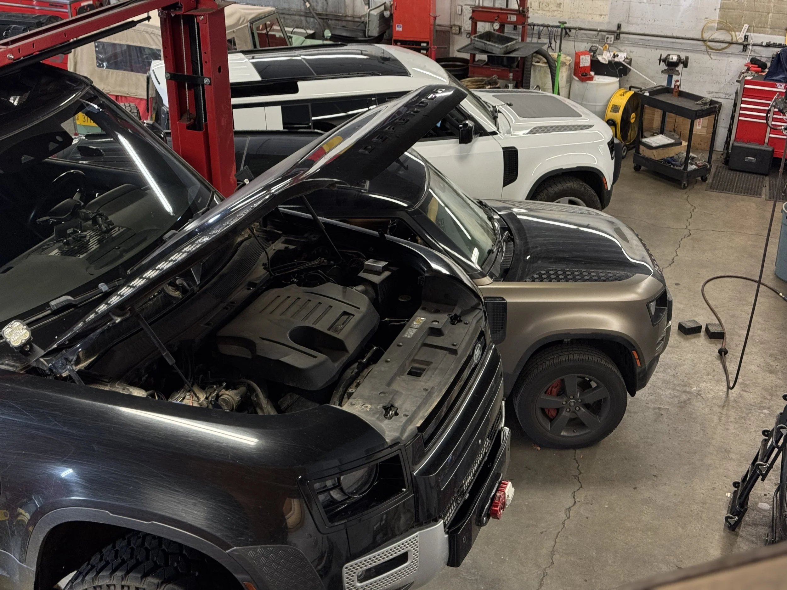 Inside an automotive workshop with multiple cars, including a black and silver car with its hood open, a white vehicle, and other cars. The background has tools, equipment, and storage cabinets.