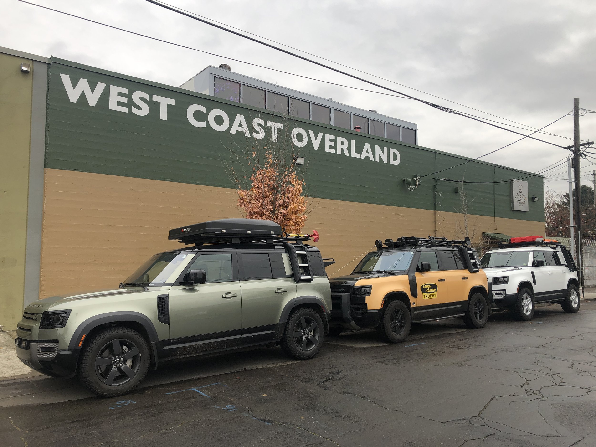 Parking lot with four Land Rover SUVs parked in front of a green and tan building with large white text that reads 'West Coast Overland' and a tree with orange leaves.