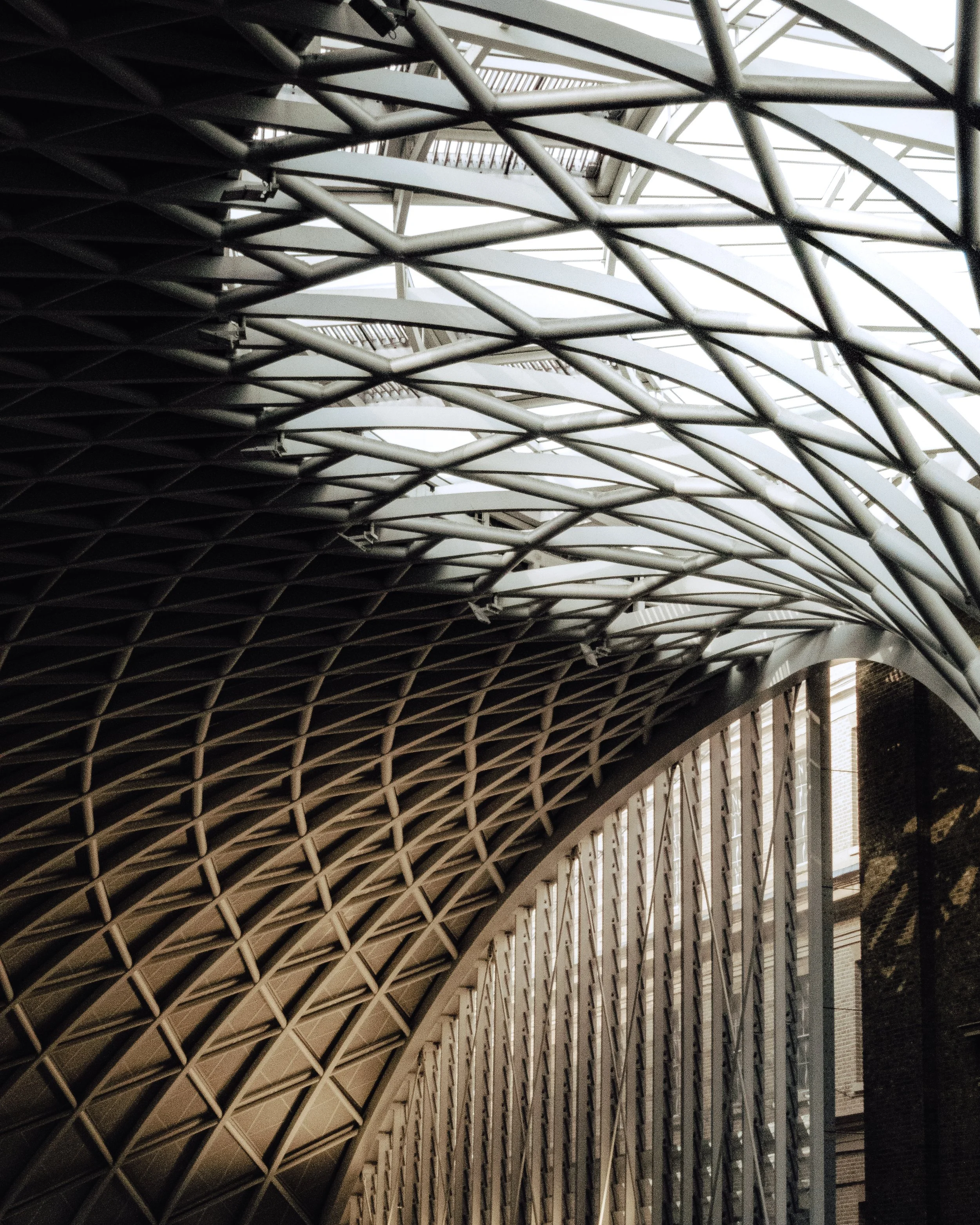 Looking up at a modern architectural structure with a curved, metallic grid pattern and glass panels in the ceiling, allowing natural light to filter through.