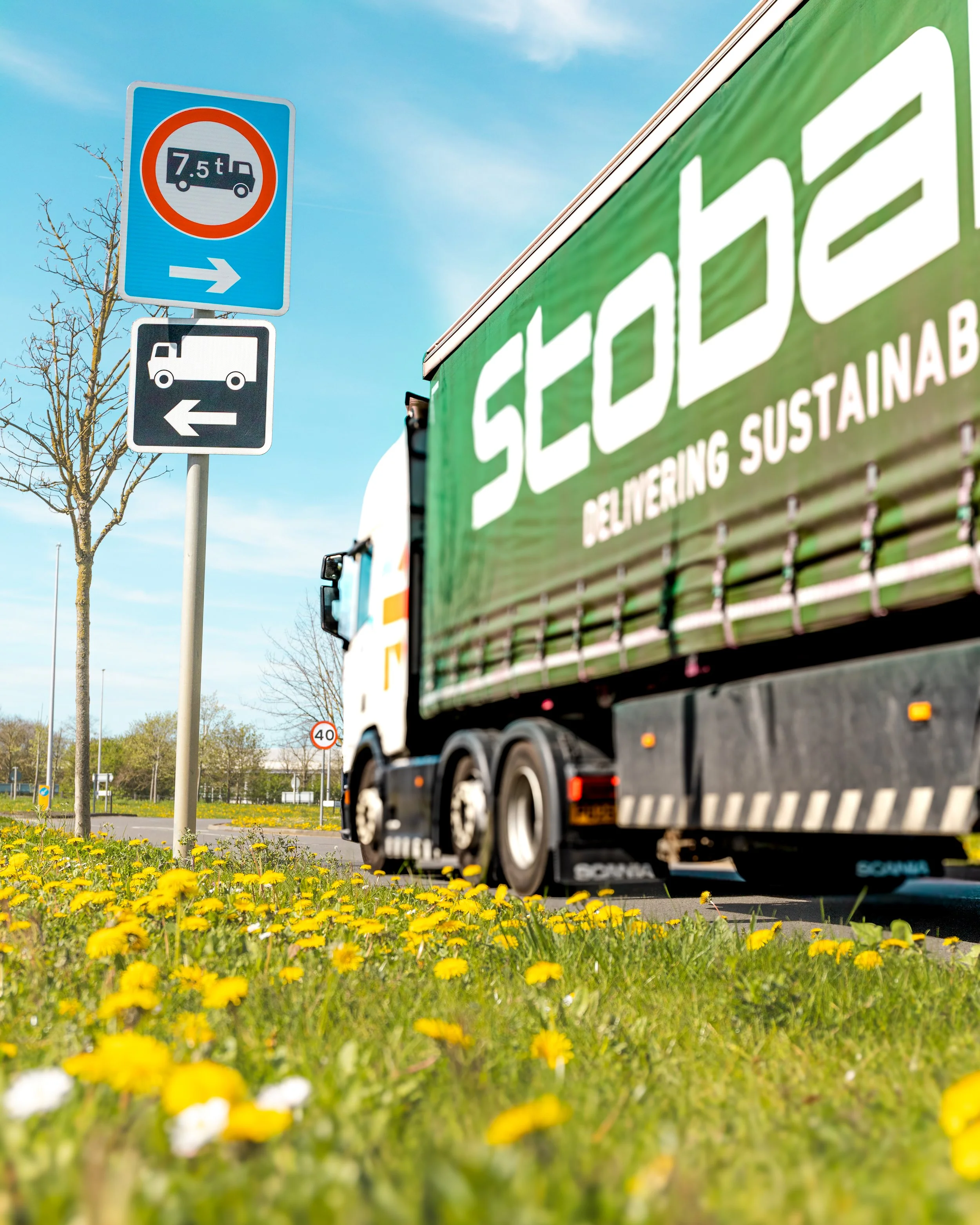 A delivery truck passes by a two-way traffic sign with arrows pointing in opposite directions, and smaller signs indicating a weight limit and speed limit, under a bright blue sky with a few clouds and a blossoming tree.