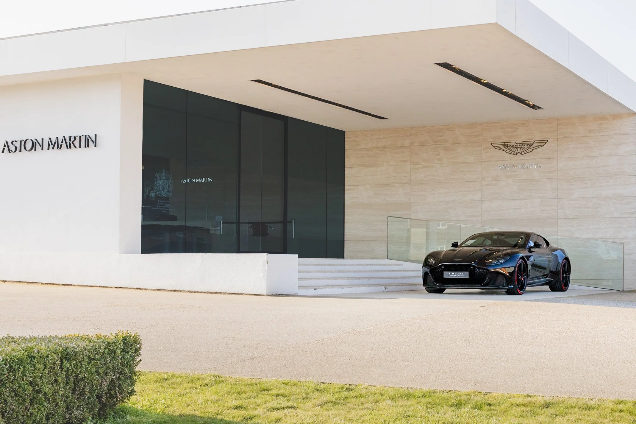 A black Aston Martin sports car parked outside a modern building with the Aston Martin logo displayed on the wall.