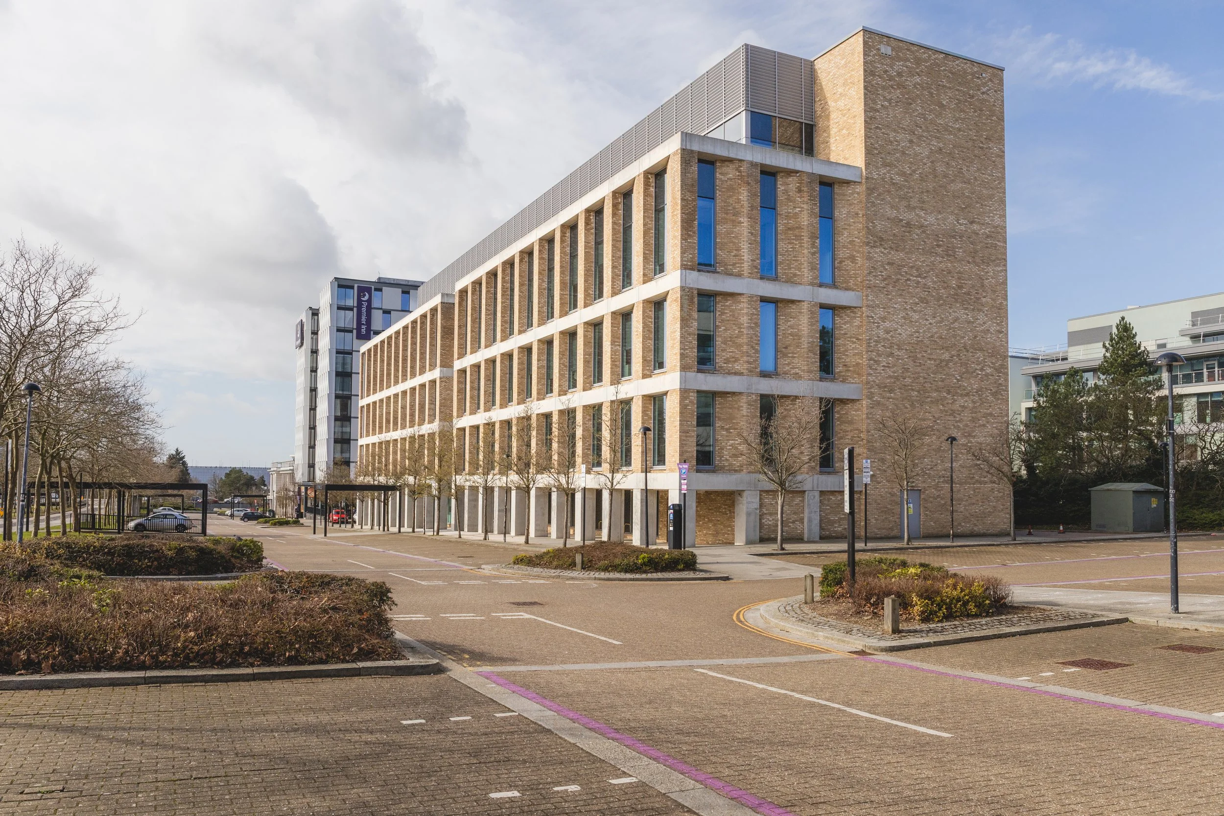 A multi-story modern building with a brick facade and large vertical windows, situated in a parking lot with marked spaces and small trees, under a partly cloudy sky.