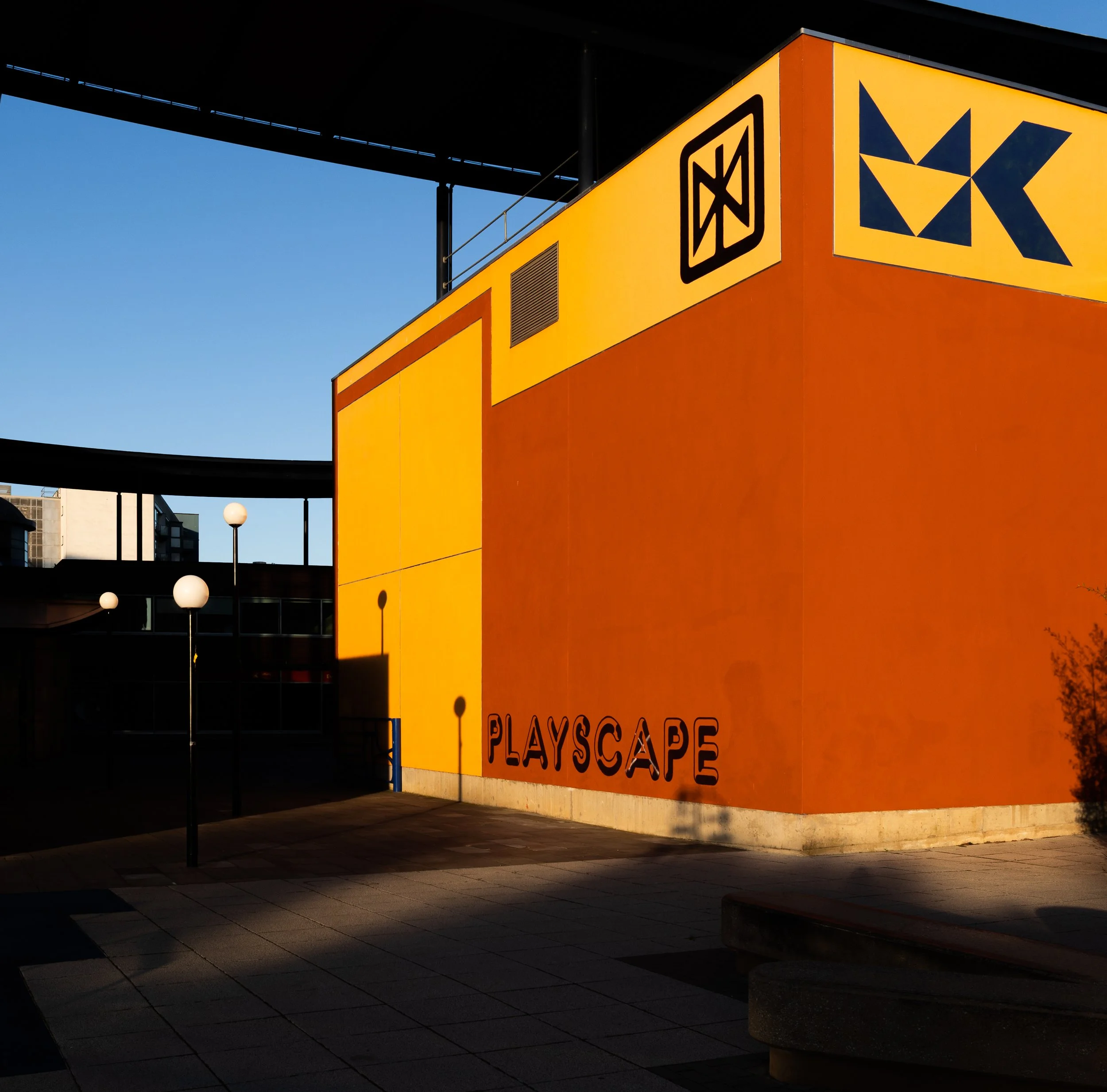 Exterior view of a brightly lit building with the sign 'Playscape' and the logo of a yellow and black starburst. The building is orange, with a black overhang above. There are street lamps and a sidewalk in front of the building, with some dark shado
