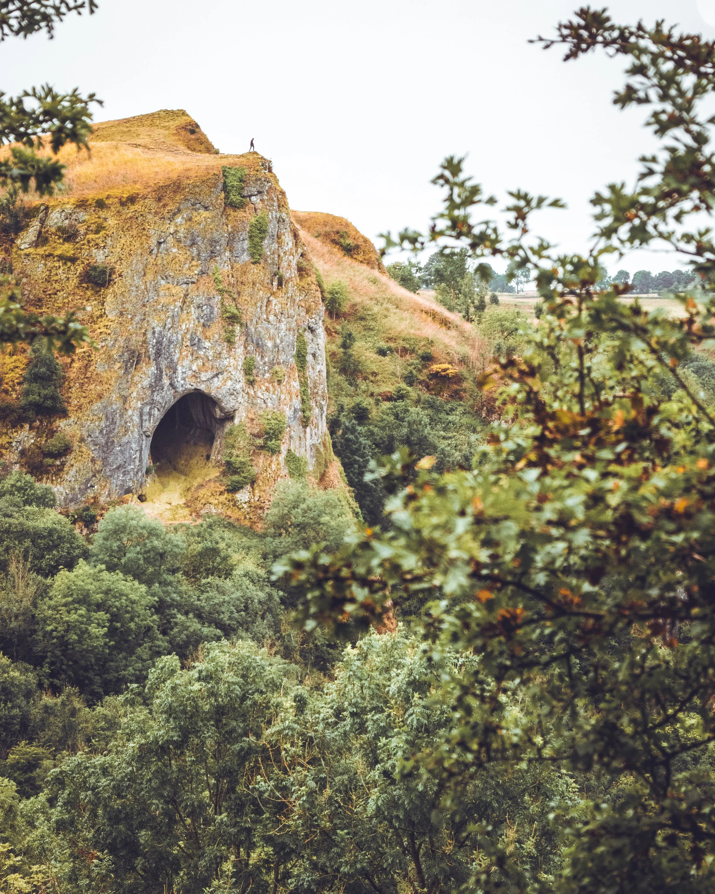 A person standing on top of a rocky hill with a large arch at its base, surrounded by trees and lush green vegetation.