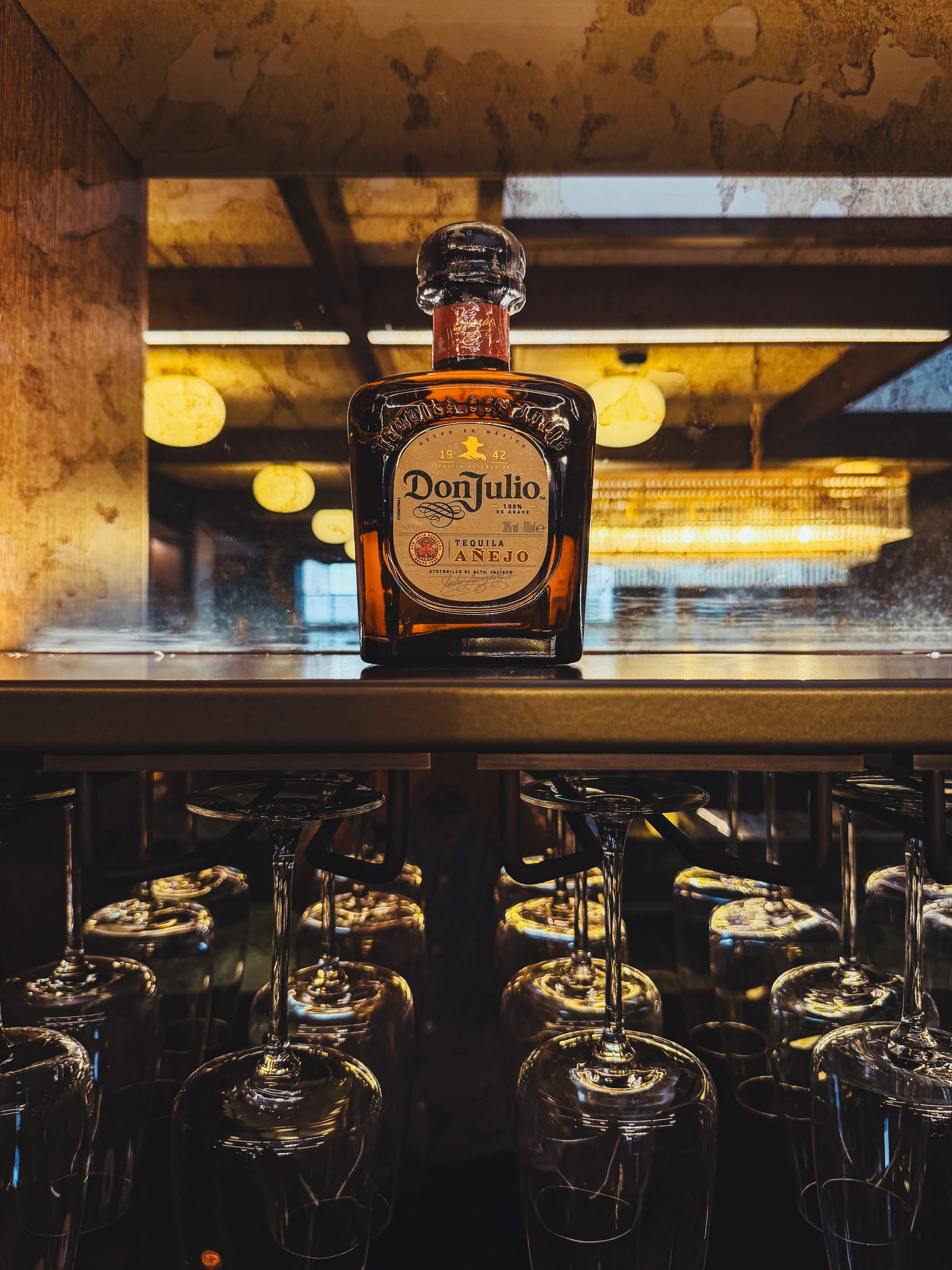 A bottle of Don Julio Añejo tequila placed on a bar counter with upside-down wine glasses hanging below.