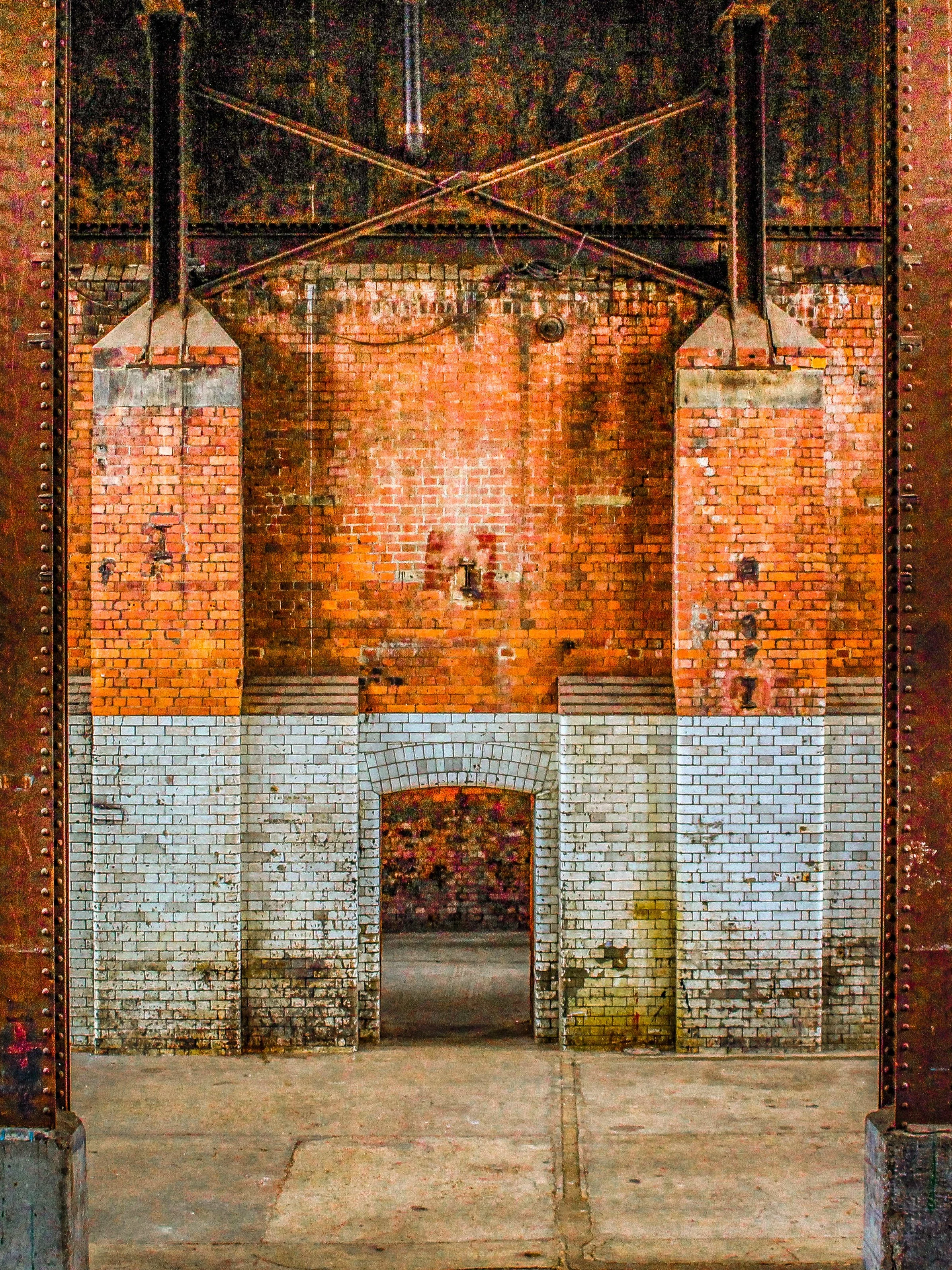 A brick wall with an arched doorway at the bottom, heavy iron hinges, and rivets with a darkened earth floor.