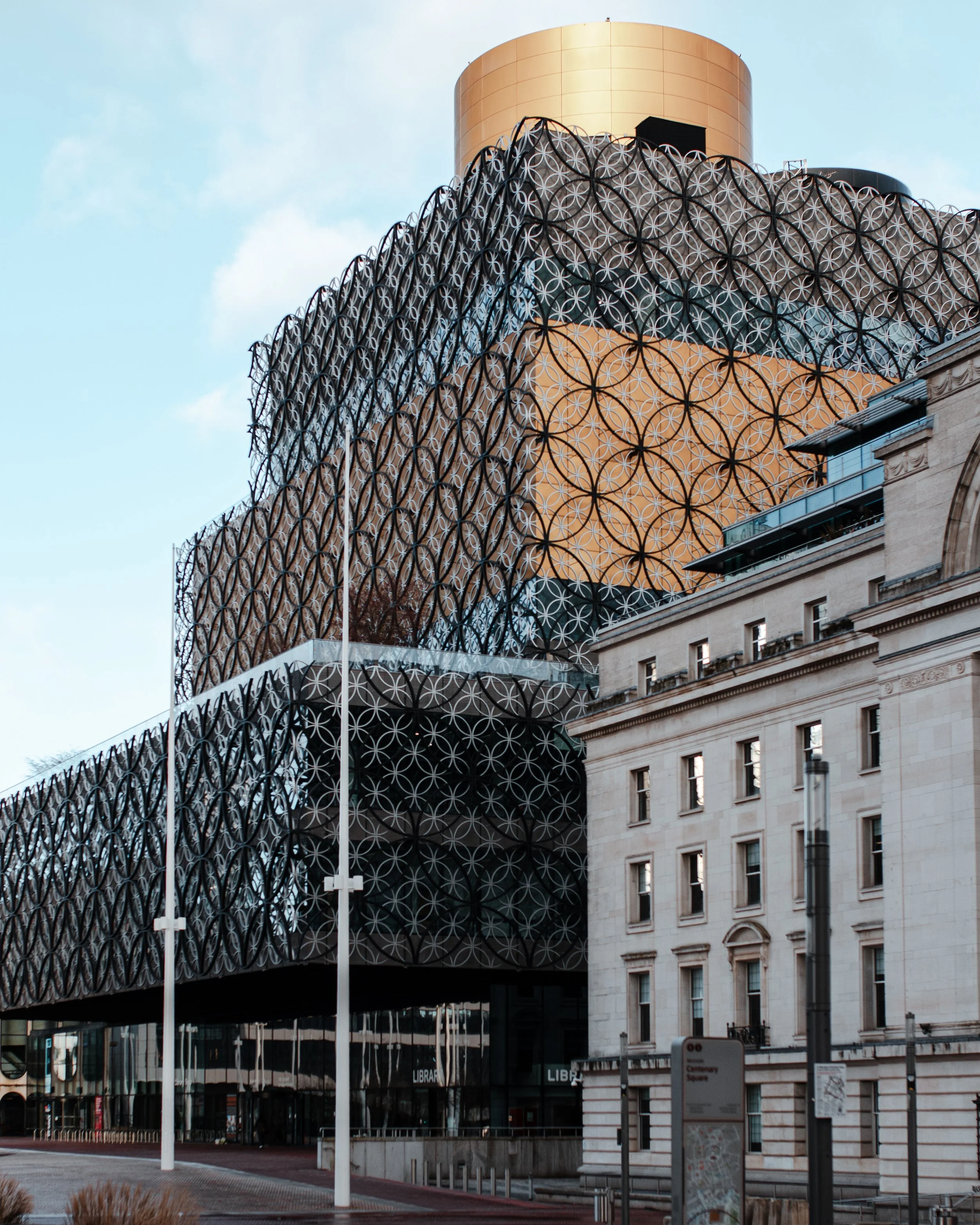 Modern building with a distinctive decorative lattice facade, a cylindrical gold-colored top, and traditional historic architecture at street level, located in an urban area.