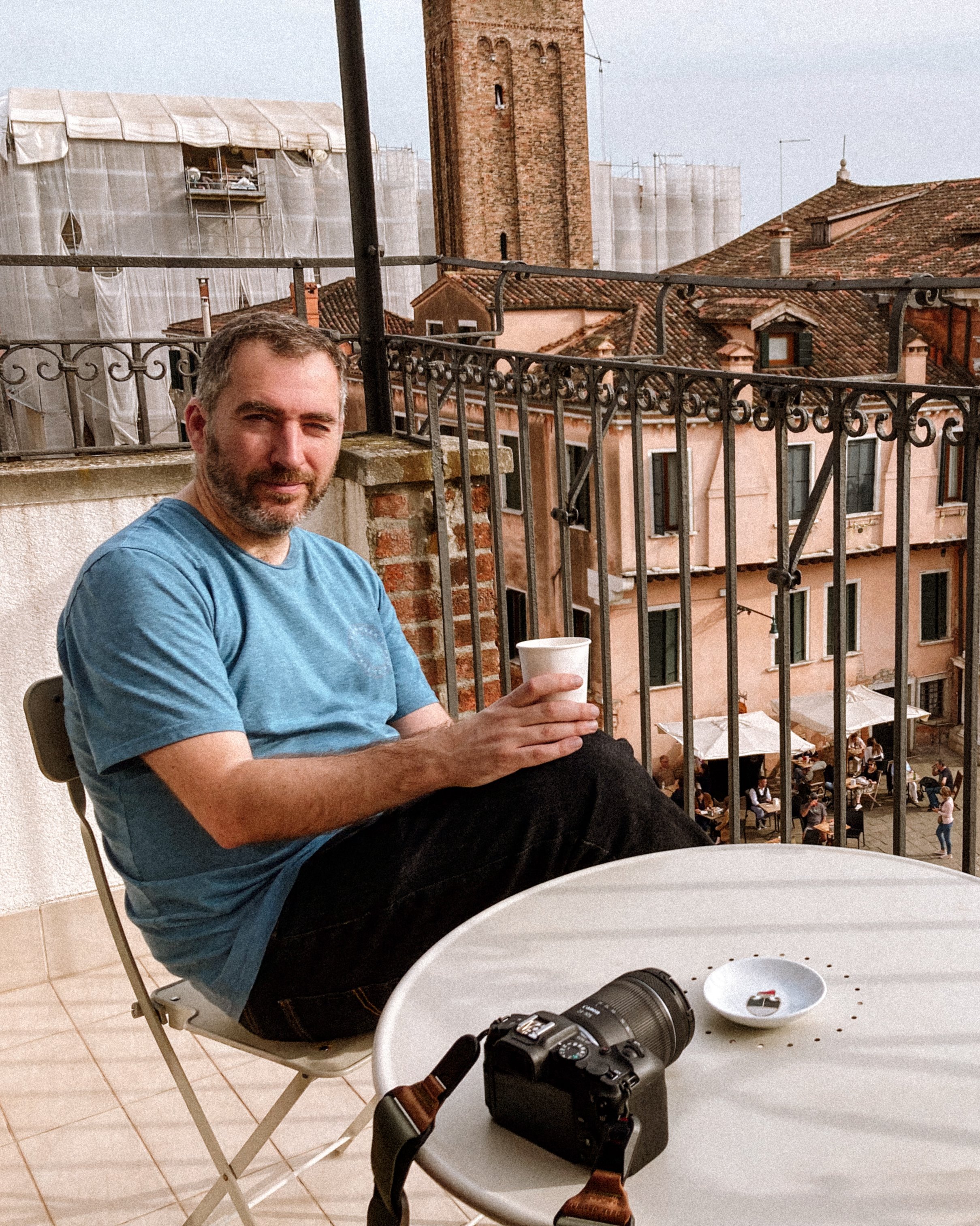 A man with a beard wearing a blue shirt sitting on a balcony holding a cup. In the foreground, there is a white table with a camera and a small white dish. The balcony has metal railings and offers a view of old buildings with tiled roofs and a tall brick tower in the background. Behind the man, there is a lens and some other objects on the table. The scene appears to be at sunset with warm lighting.