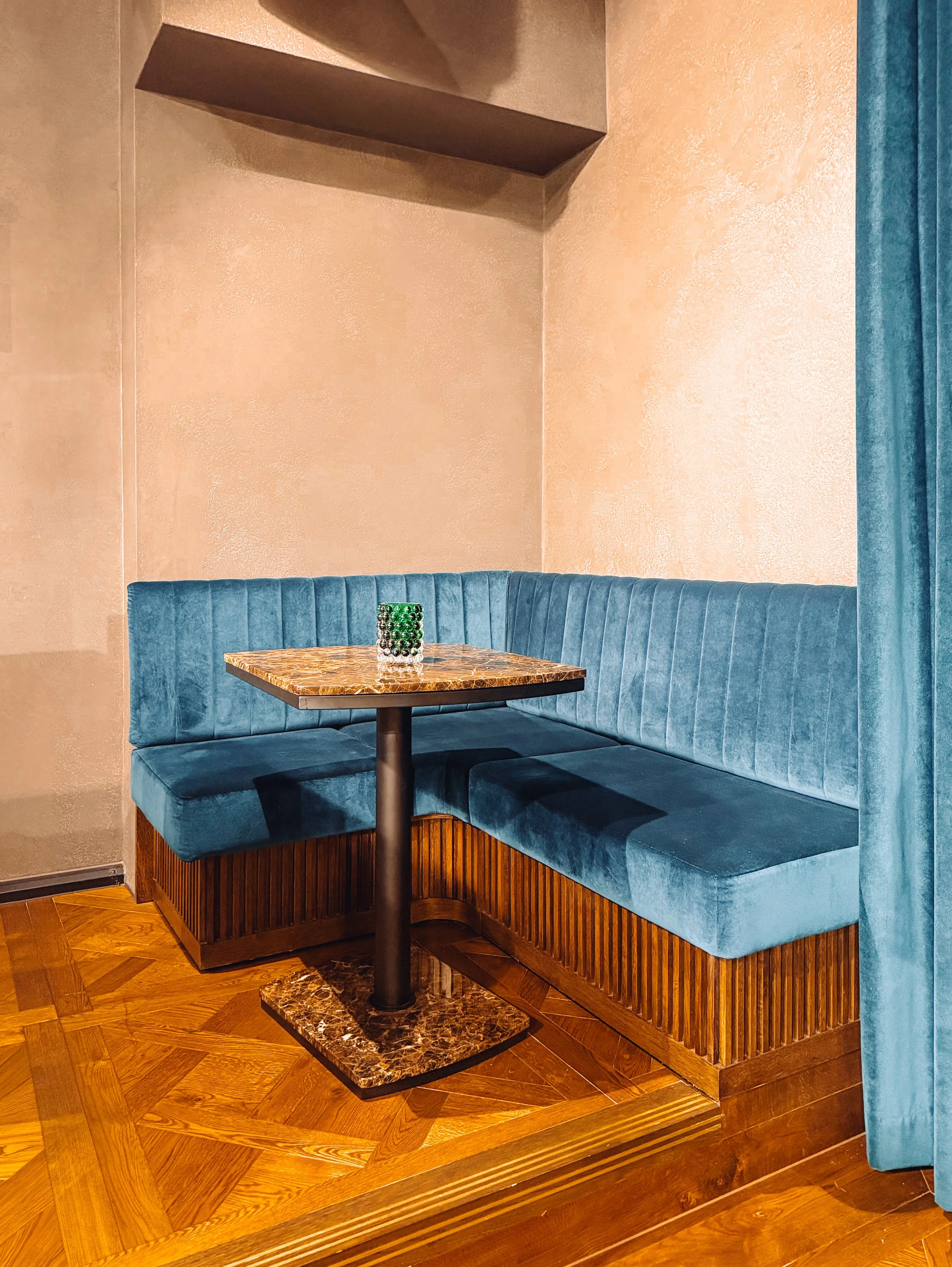 A corner booth with blue upholstered seating and a small marble-top table with a decorative object on it, in a room with peach-colored textured walls and wooden flooring.