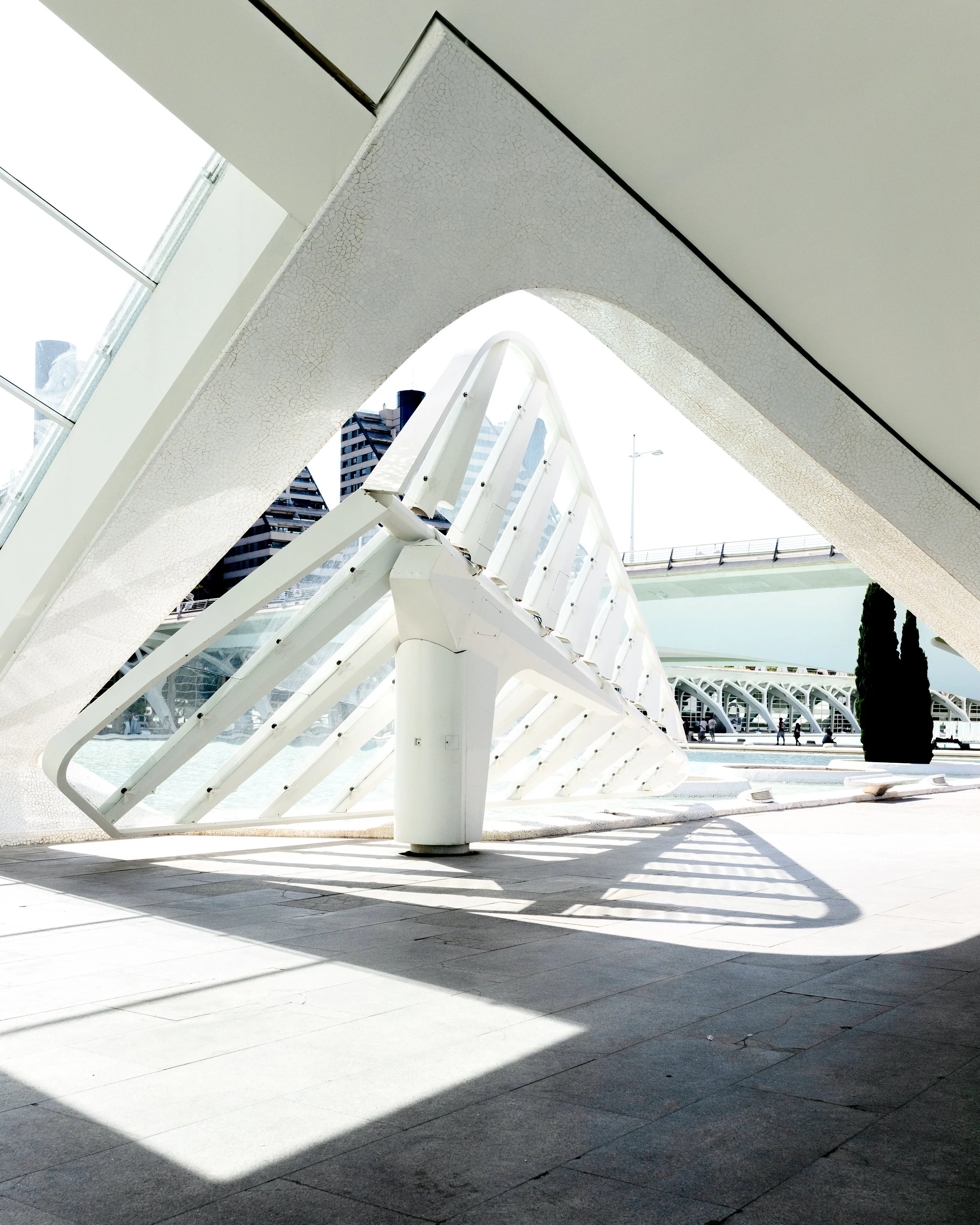 Modern architectural structure with sweeping white arches and open walkways in an urban setting, with tall buildings in the background and shadows cast on the ground.