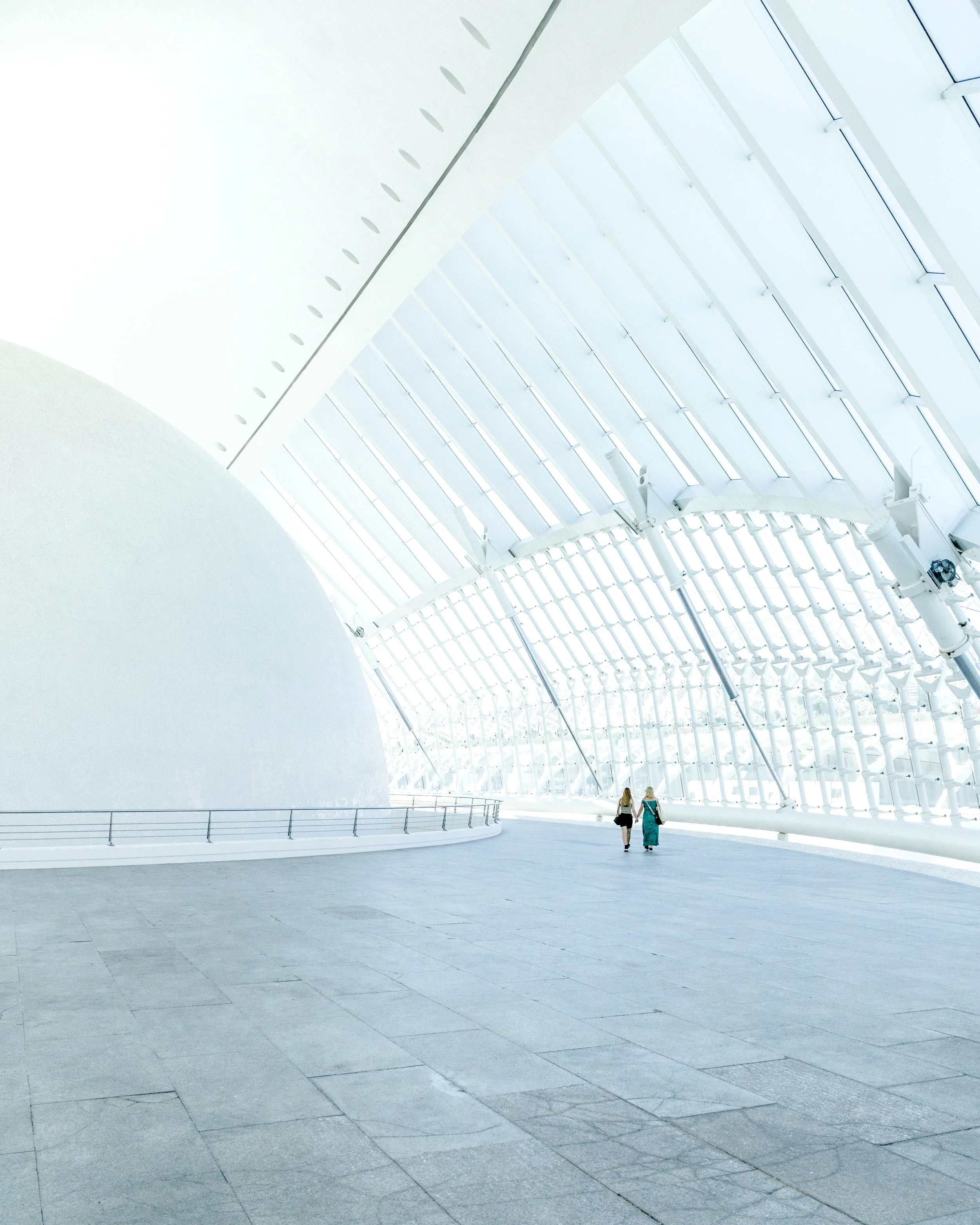 Modern architectural interior with curved white walls, large glass windows, and a high ceiling, with two women walking together.