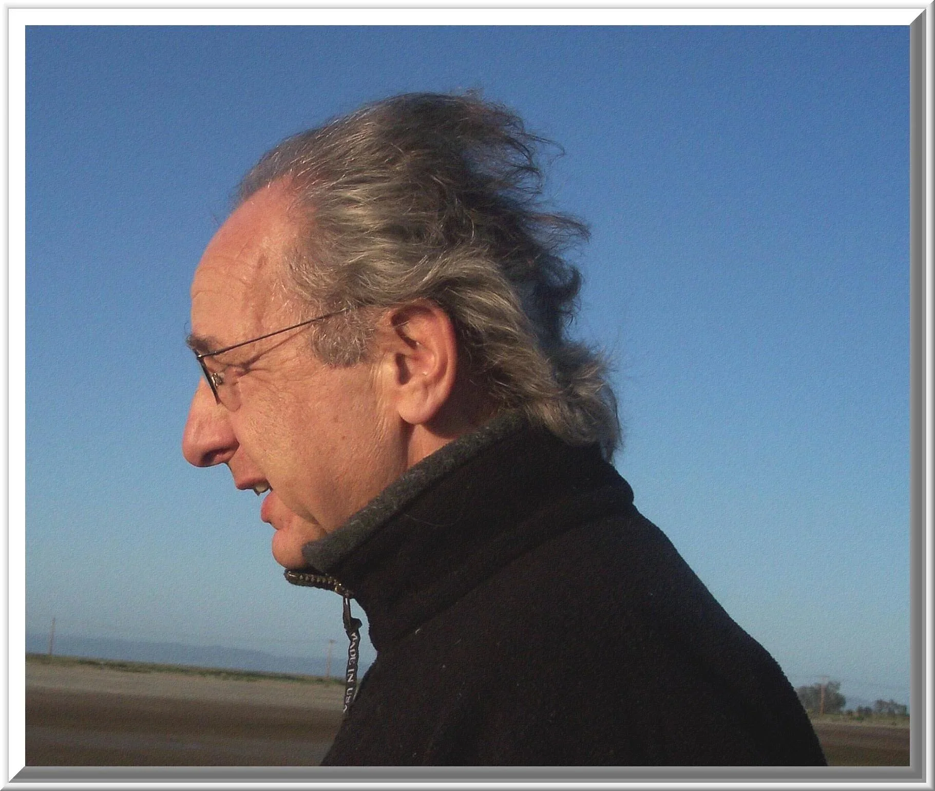 A close-up side profile of a middle-aged man with gray hair, glasses, smiling, wearing a black jacket with a zipper, outdoors against a clear blue sky, with a blurred landscape background.