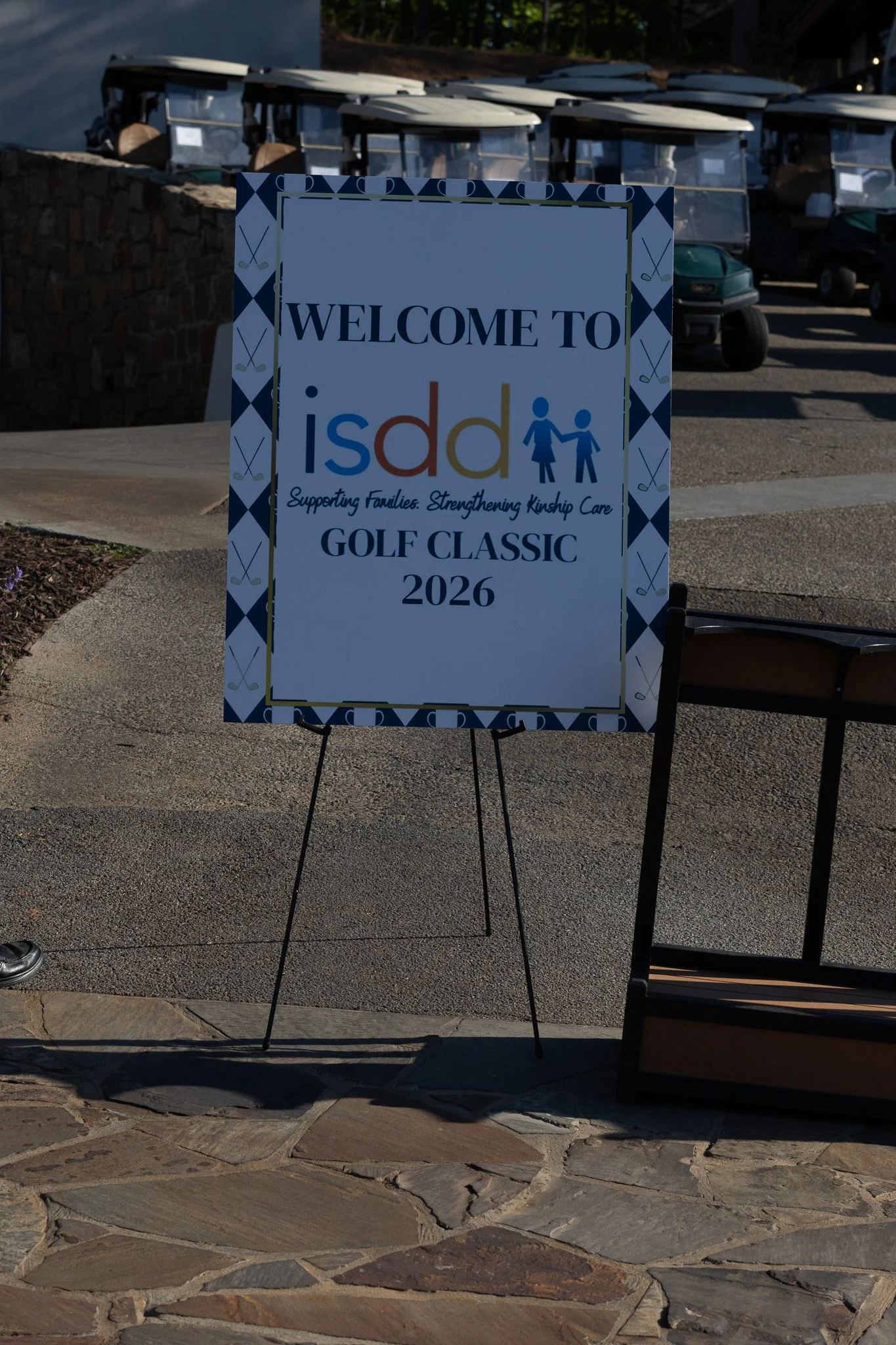 A sign welcoming people to the ISDA Golf Classic 2026 with the slogan supporting families and strengthening kinship care, surrounded by golf-themed decor, with golf carts parked in the background.