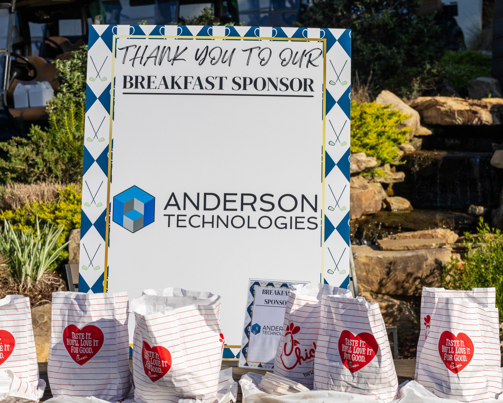 A thank you sign for a breakfast sponsor, Anderson Technologies, in an outdoor setting with plants and a water feature in the background. There are paper lunch bags in the foreground, some with Chick-fil-A logos and messages.